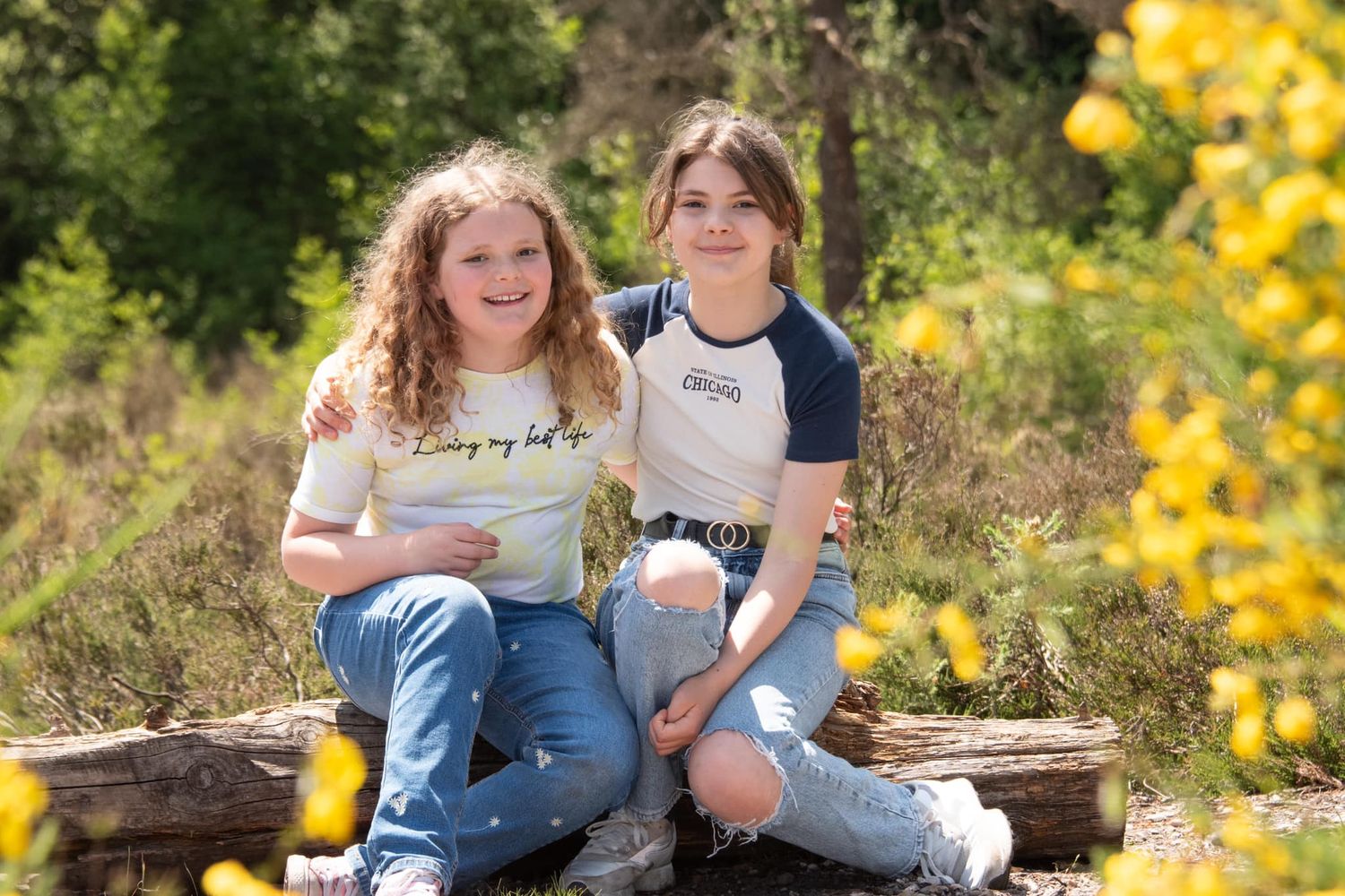 Two smiling teenage girls sitting on a log surrounded by bright yellow wildflowers in a sunny outdoor setting.