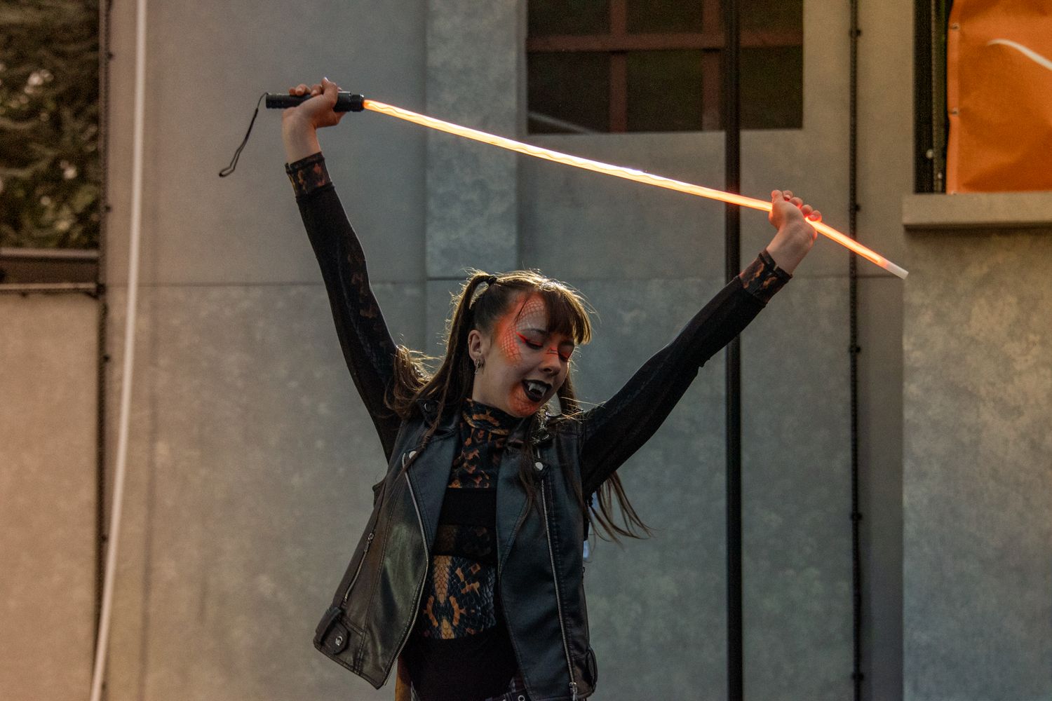 Female performer in black leather jacket triumphantly raising two drumsticks above her head on an outdoor stage.