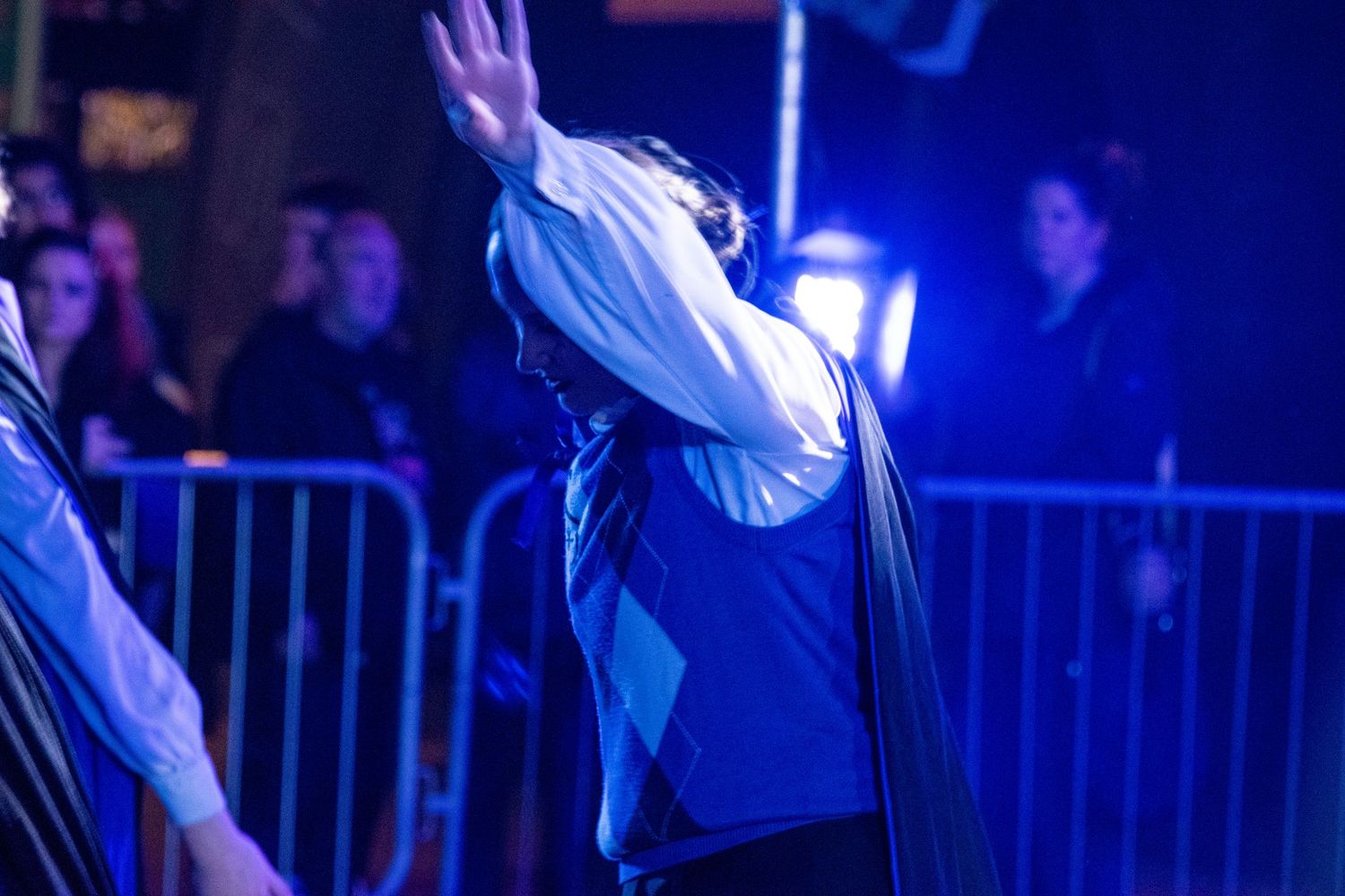 Performer in white shirt and dark vest raises arm dramatically under blue stage lighting.