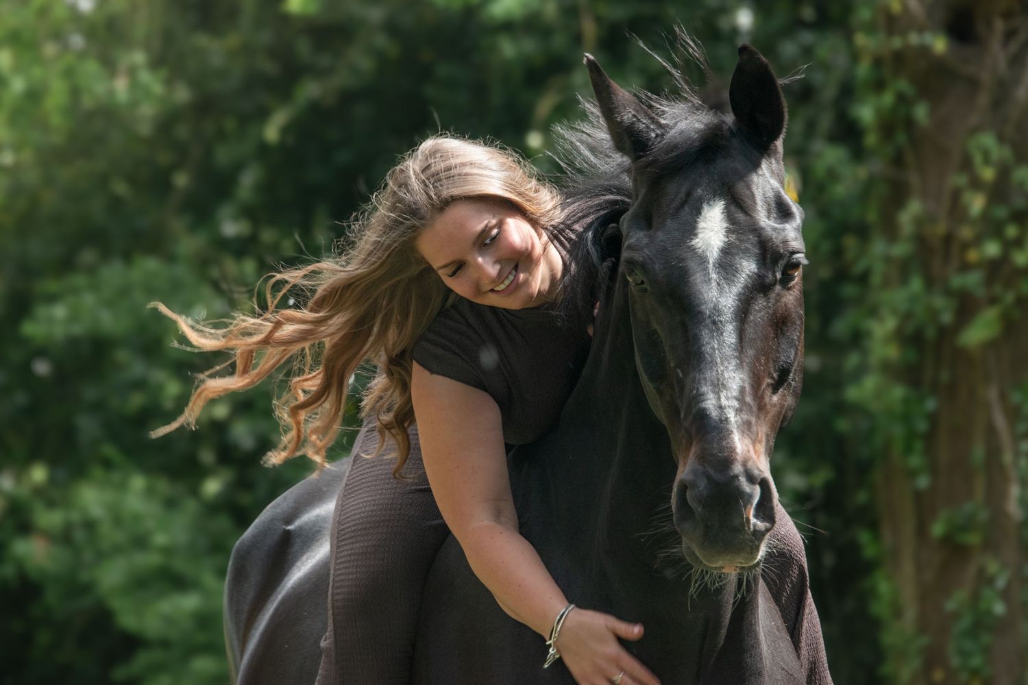Blonde woman smiling and leaning forward while riding a black horse through a lush green forest.