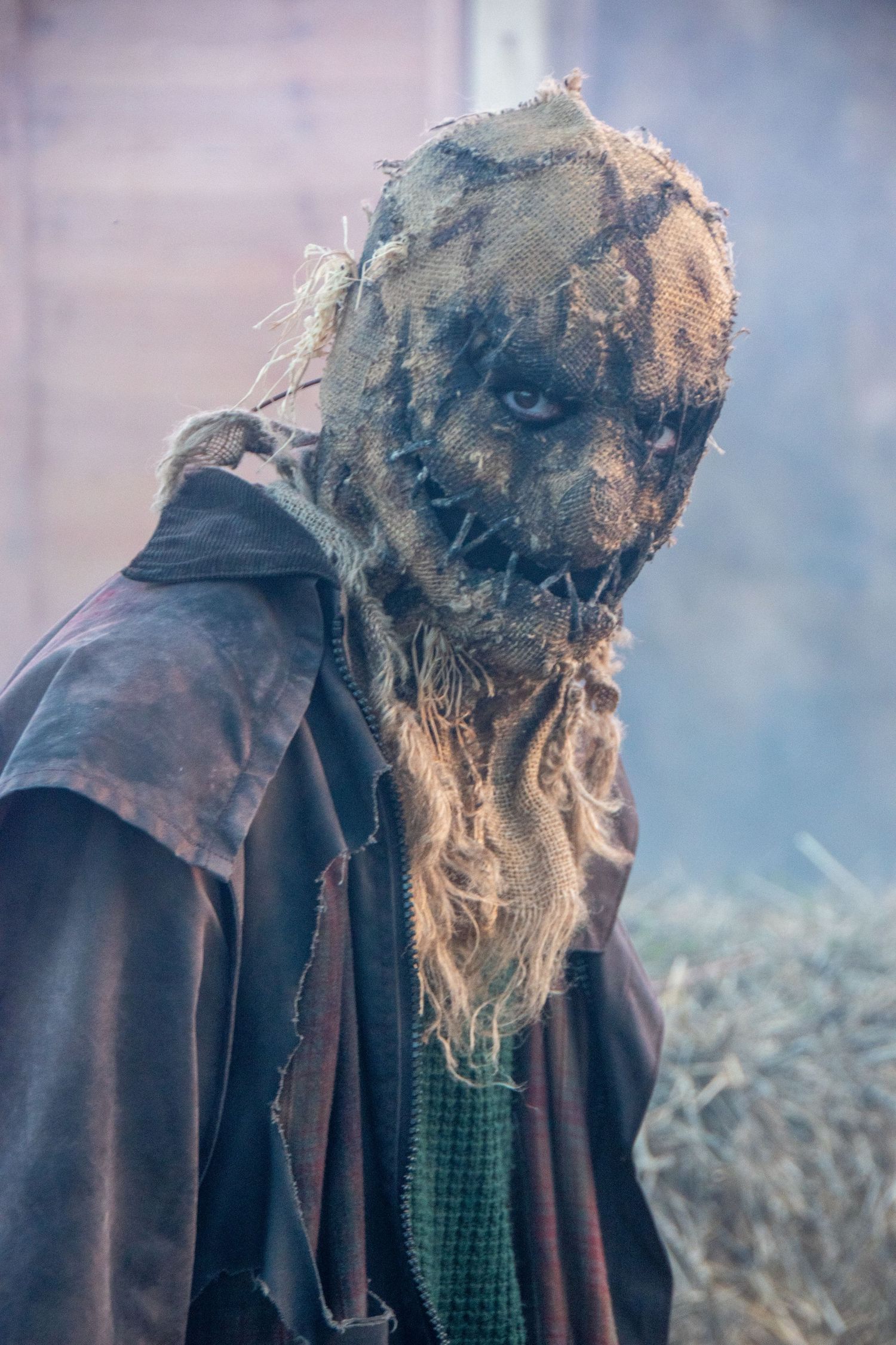 Creepy scarecrow Halloween costume with burlap mask, straw details, and tattered brown coat near hay bales.