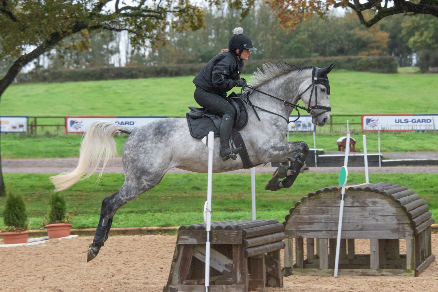 A rider on a dapple grey horse jumps over a fence obstacle in an equestrian competition.