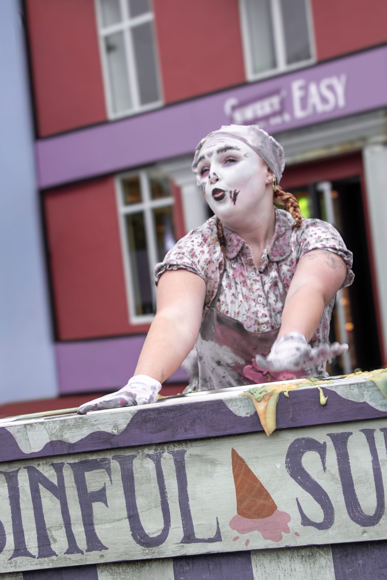 Female mime performer in white face paint leaning over a colorful street cart outdoors.