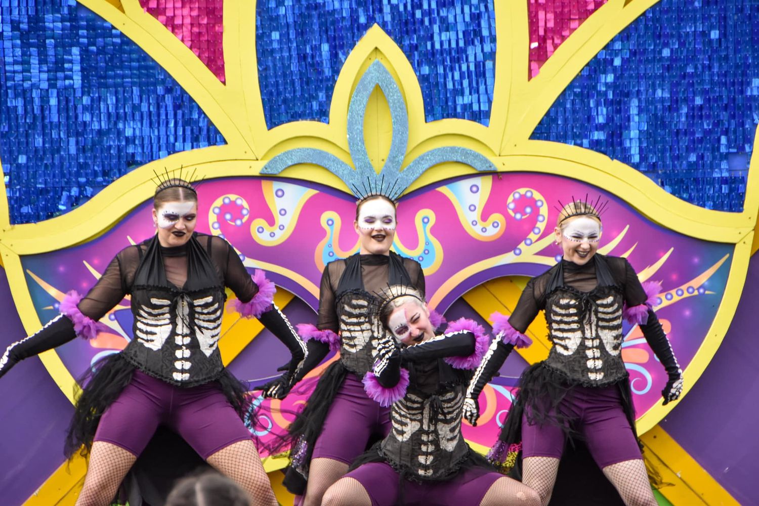Four performers in skeleton costumes posing in front of a vibrant decorative carnival backdrop.
