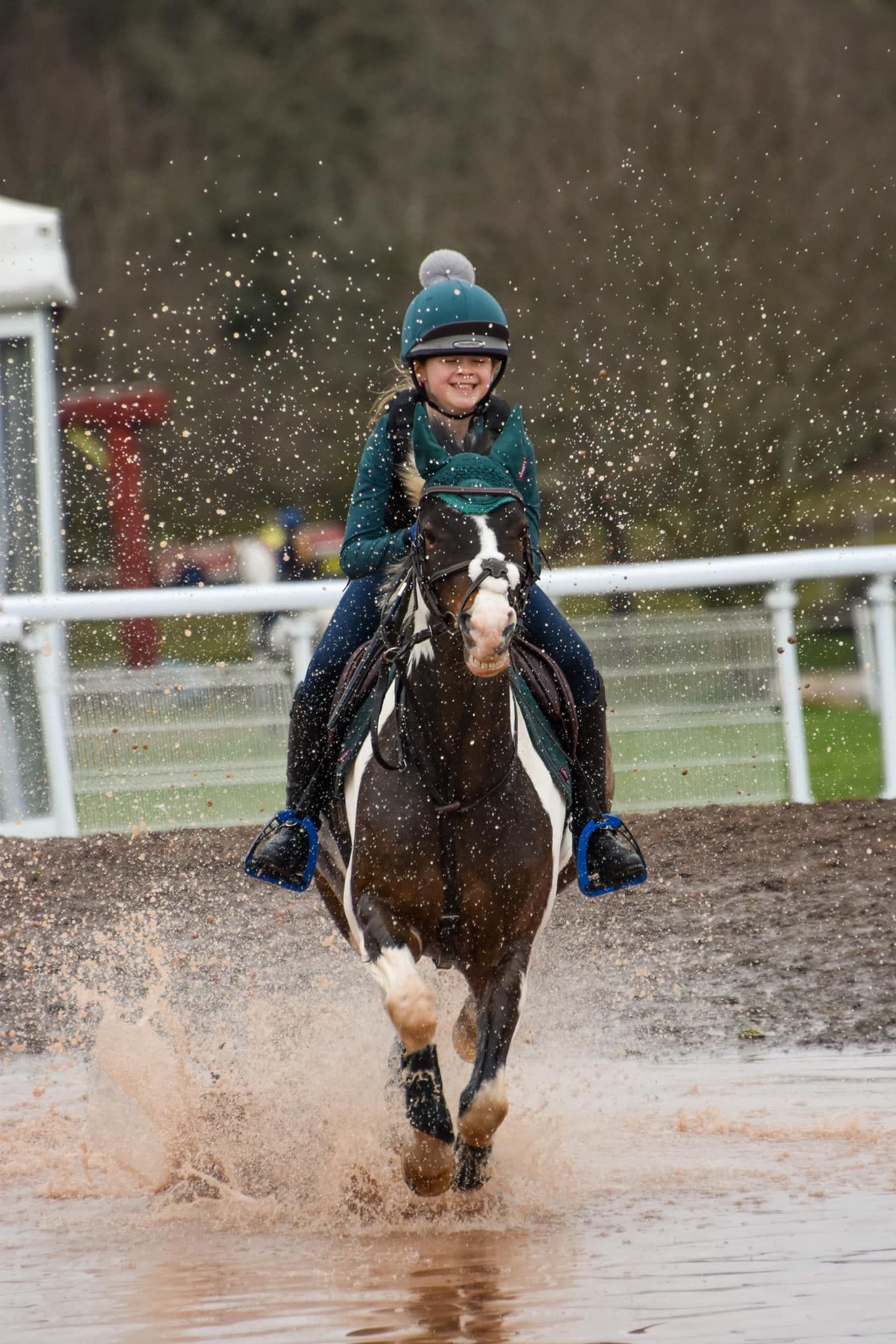 Smiling rider on a pinto horse splashing through water during a cross-country equestrian event.