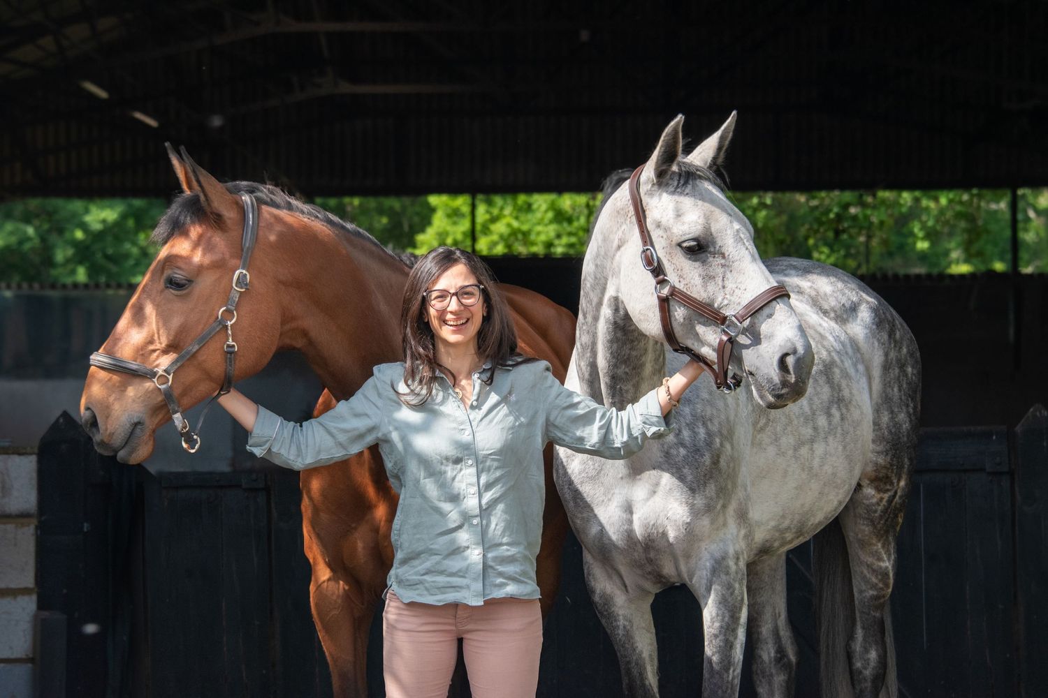 Smiling woman in a light blue shirt standing between a chestnut and a grey horse outside a stable.