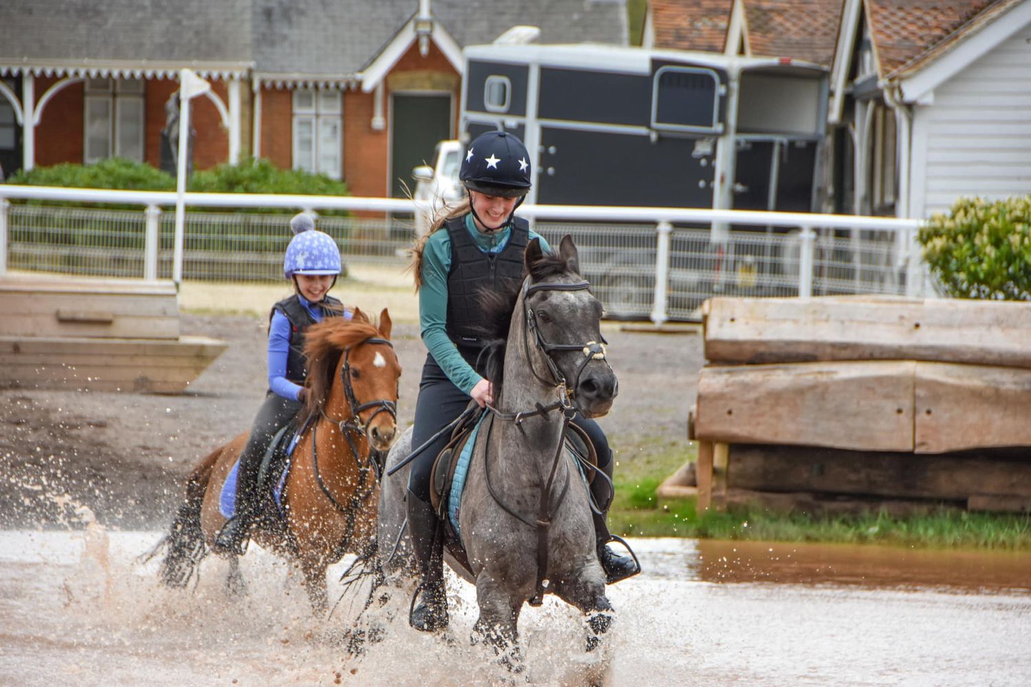 Two riders on horses splashing through water during equestrian event.