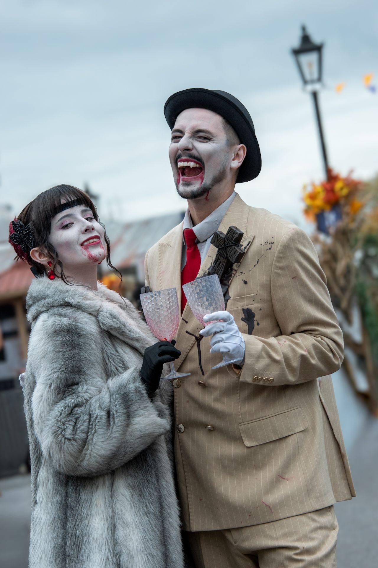 Couple in zombie Halloween costumes with face paint holding champagne glasses outdoors.