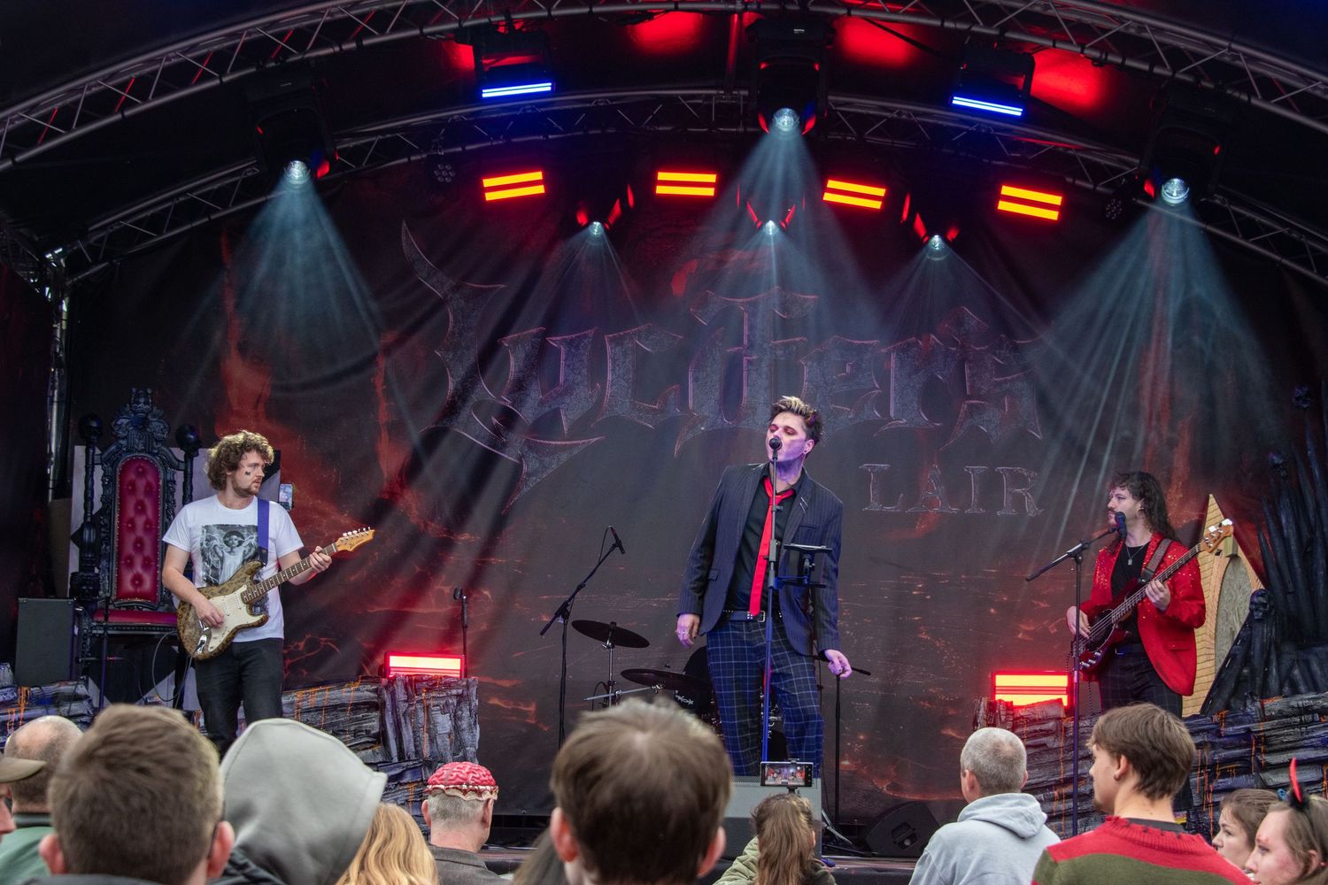 Rock band performing on an outdoor stage with colorful lights and crowd in foreground.