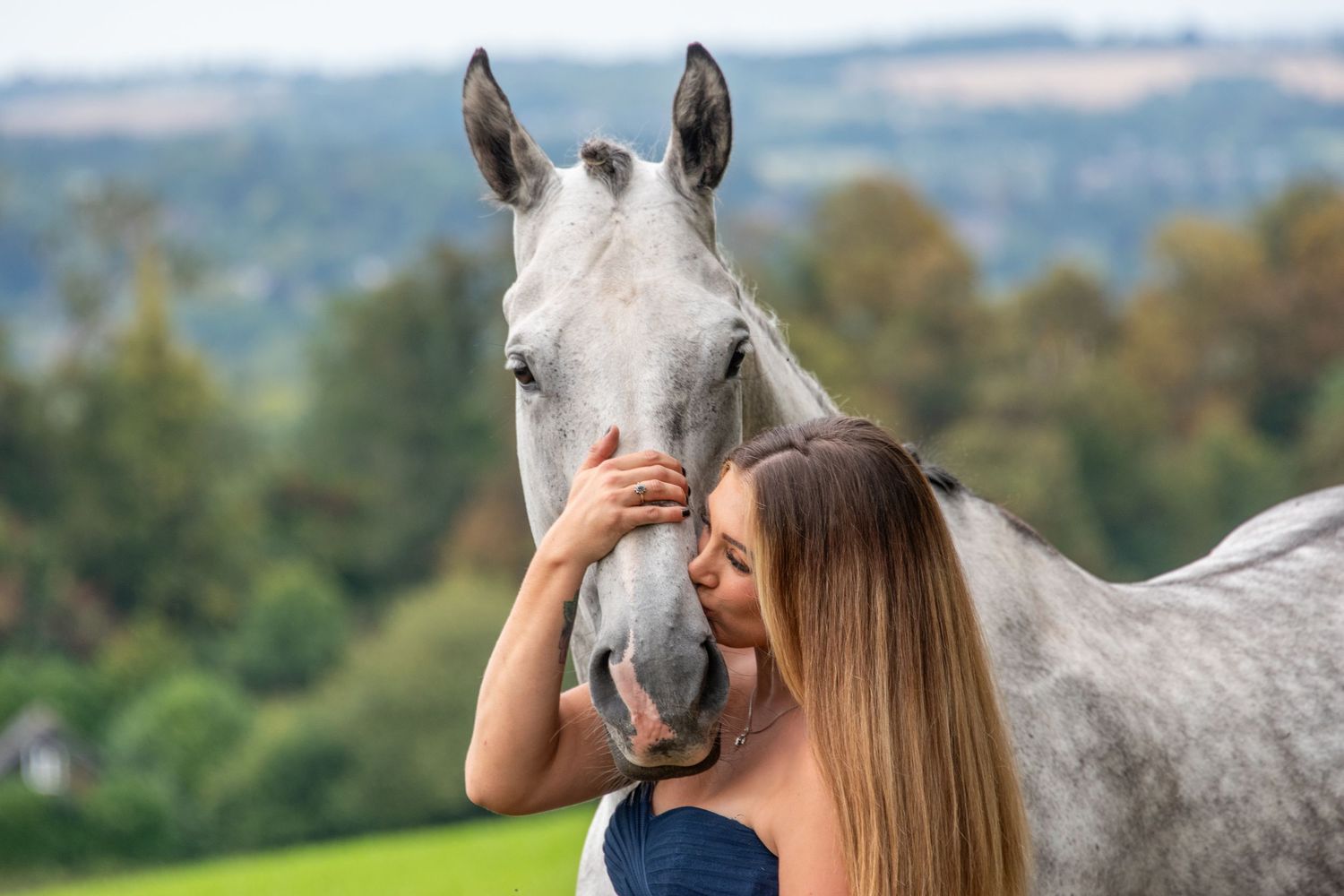 A woman with long hair tenderly embraces a grey horse's face in a scenic green landscape.