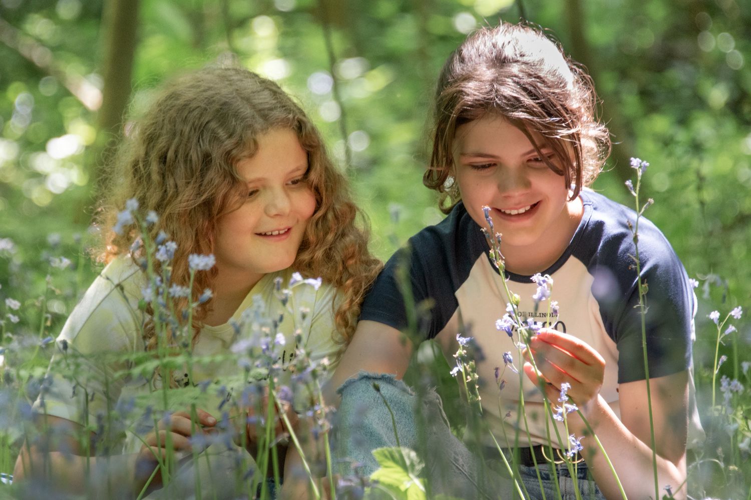 Two children smiling and examining wildflowers together in a lush green forest setting.