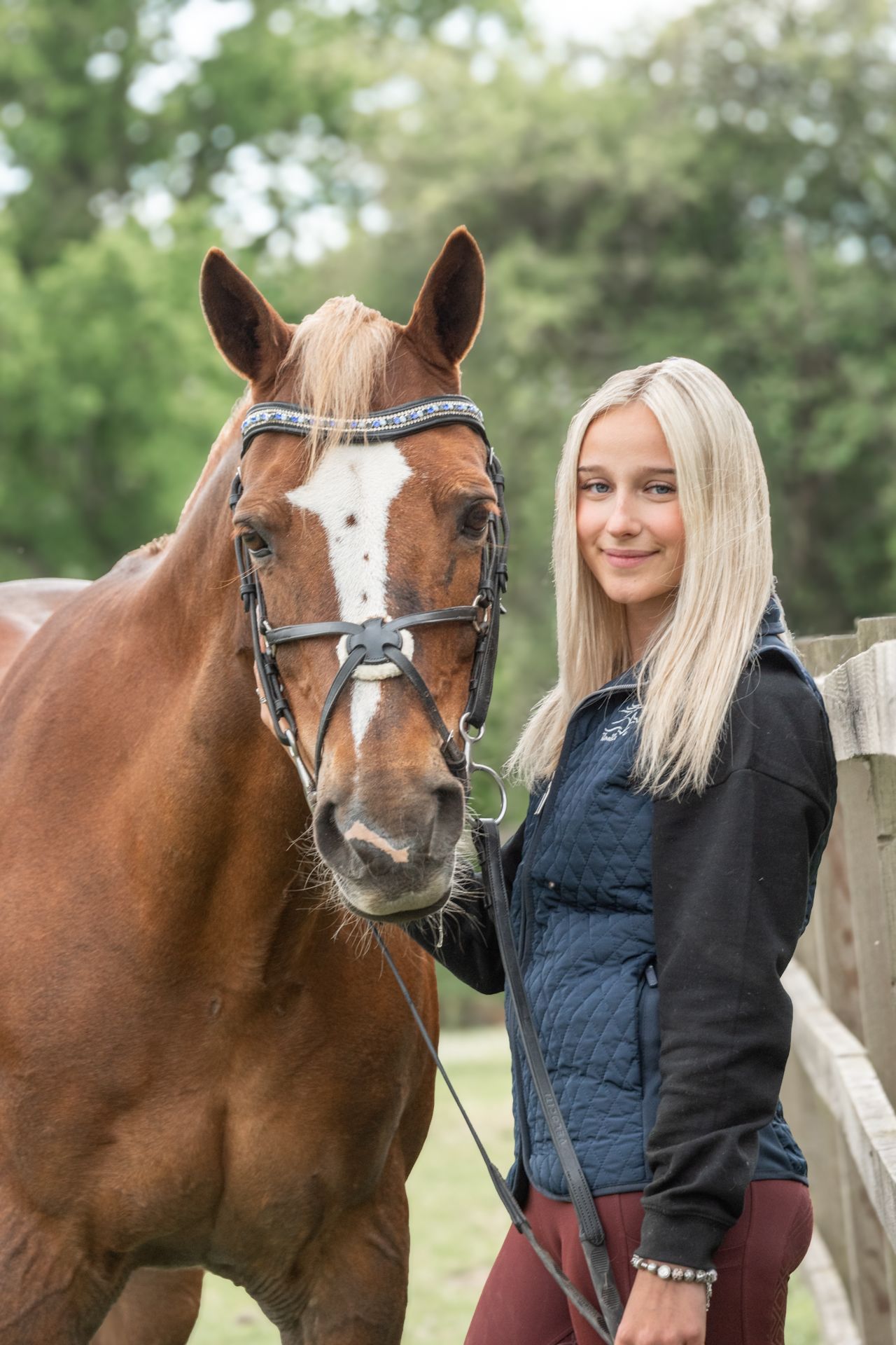 Young blonde woman smiling beside a chestnut horse with a white blaze, wearing a bridle near a wooden fence.