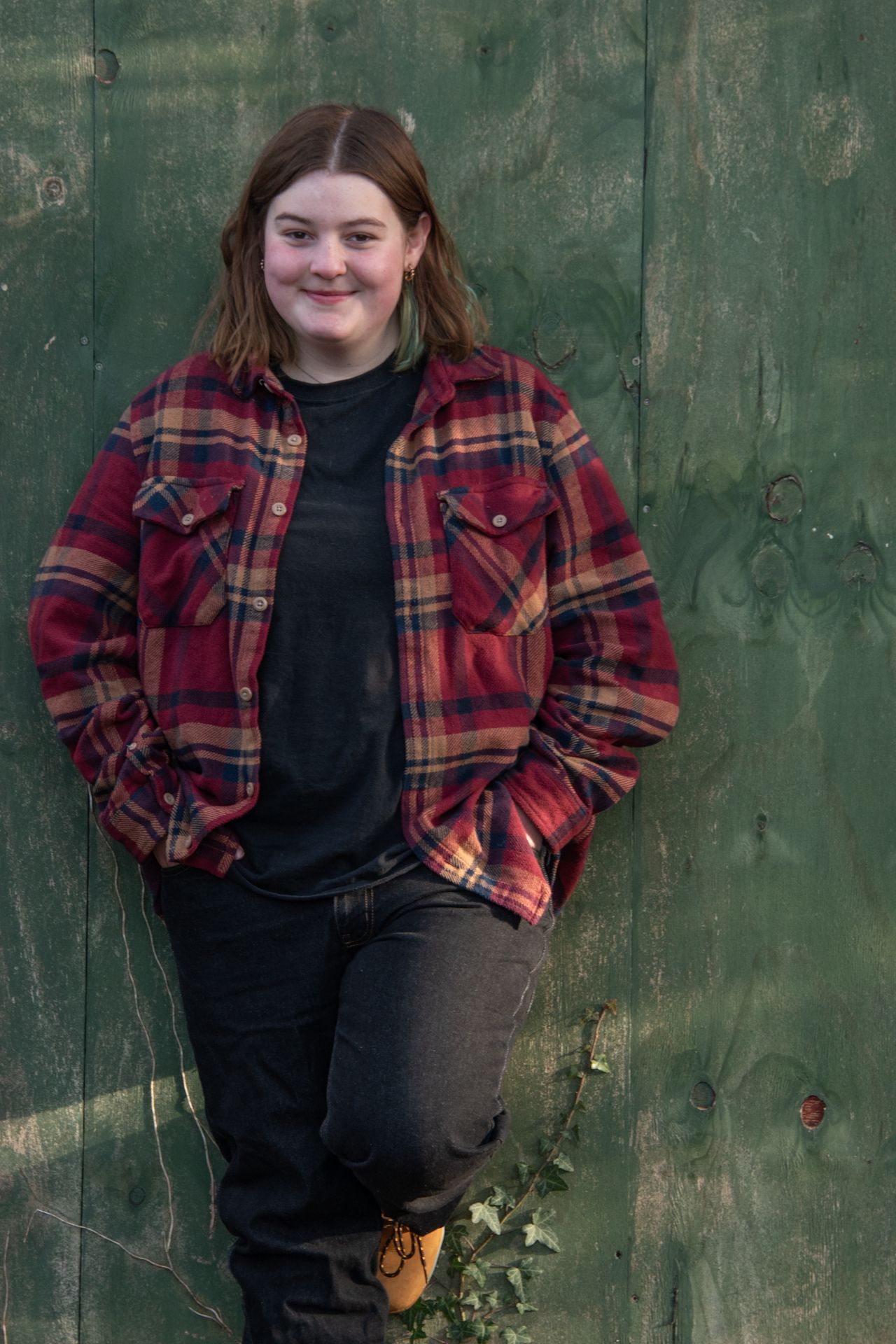 Young woman in red plaid flannel shirt leaning against green wooden wall, smiling outdoors.