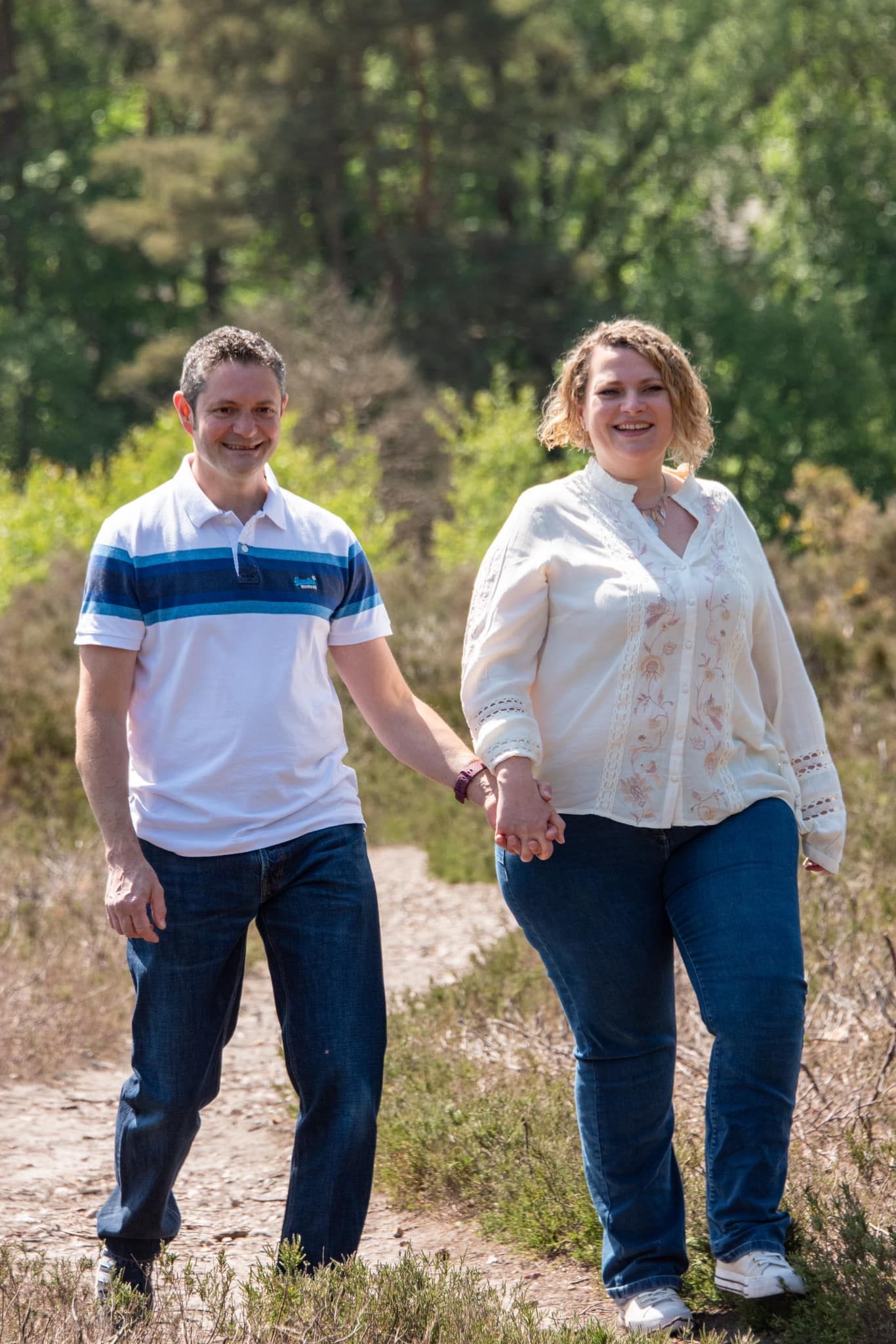 Smiling couple holding hands while walking along a sunny nature trail surrounded by green trees and shrubs.