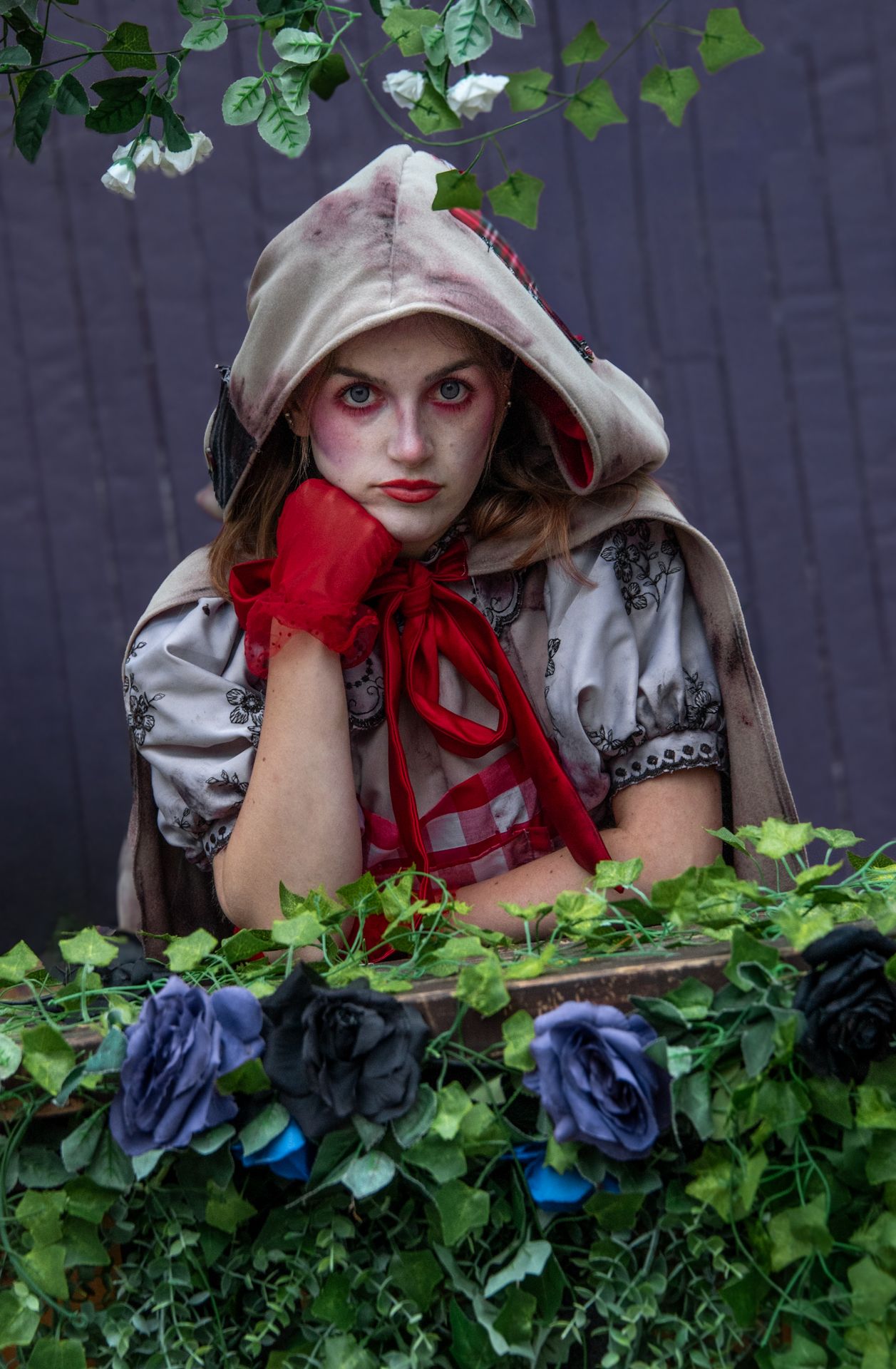 Girl in dark Little Red Riding Hood Halloween costume with dramatic makeup posing among black roses and ivy.