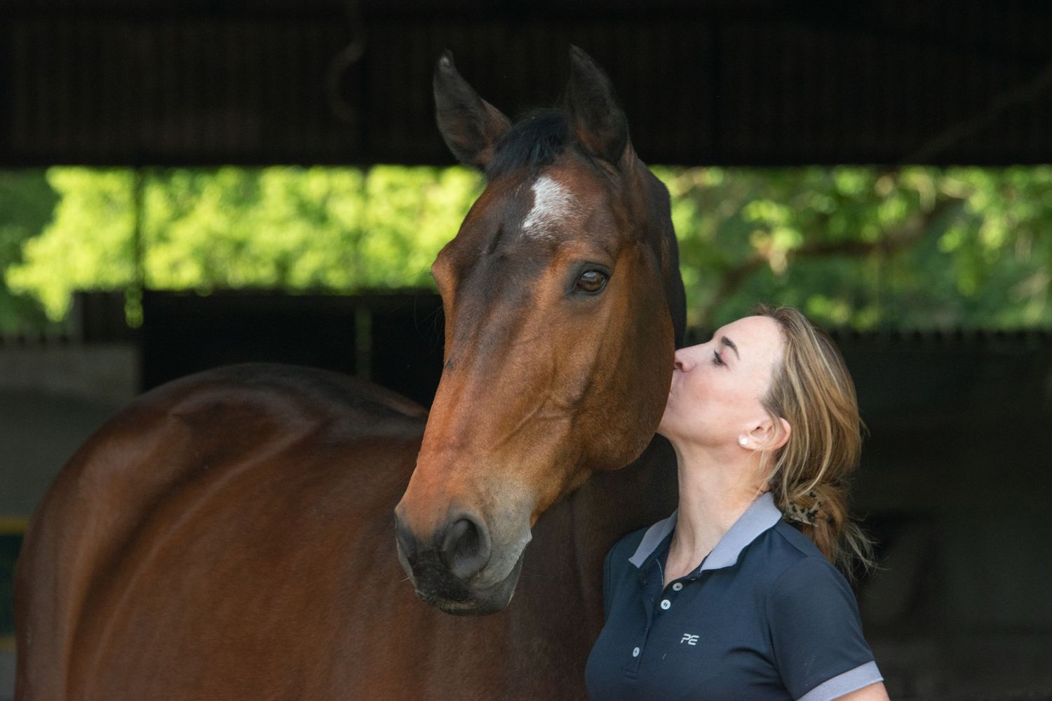 Woman in navy polo shirt sharing a tender moment kissing a bay horse in a stable setting.