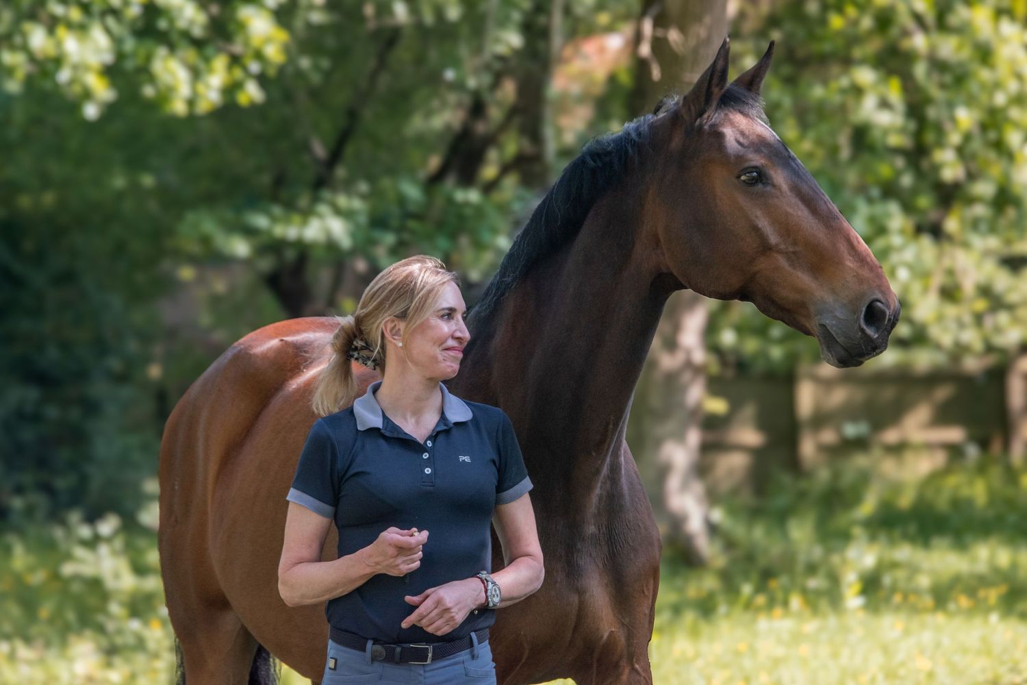 Blonde woman in a navy polo shirt walking alongside a bay horse in a sunny green meadow.