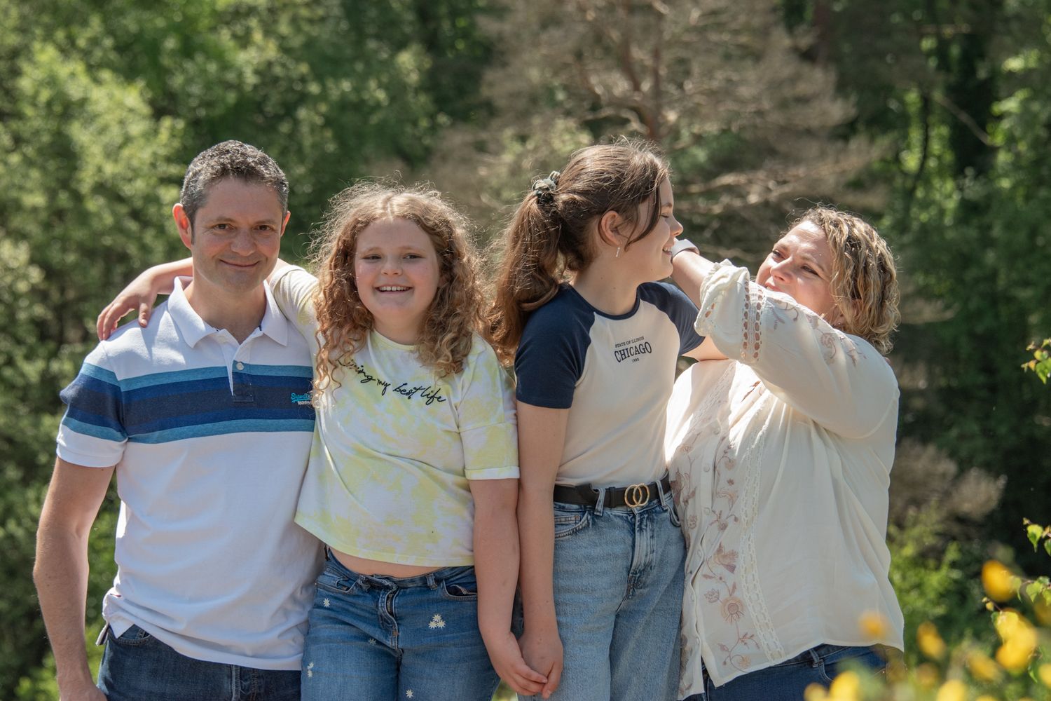 A happy family of four poses outdoors surrounded by lush green trees on a sunny day.