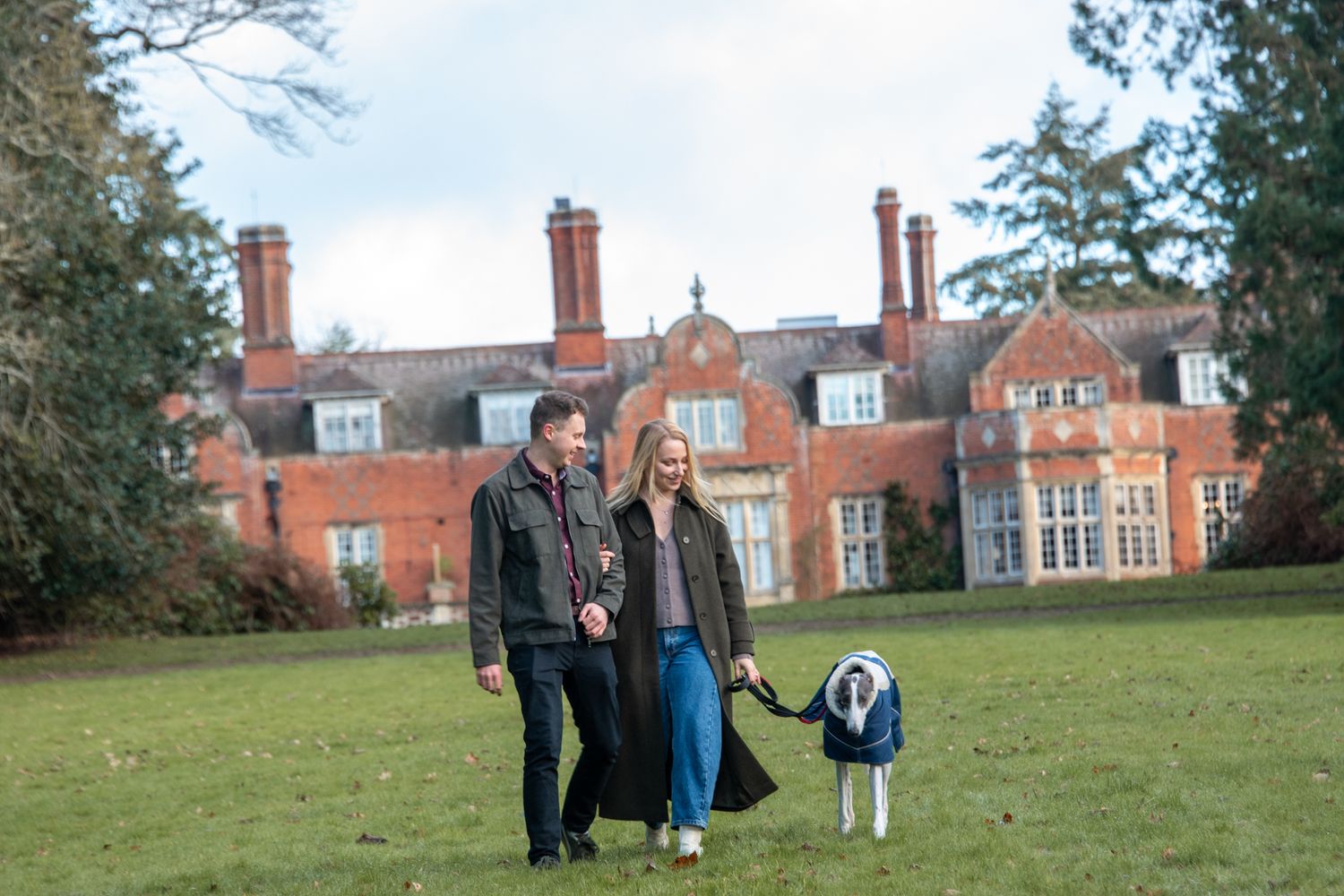 Engaged couple walking their whippet dog on a lawn in front of a grand red brick country estate.