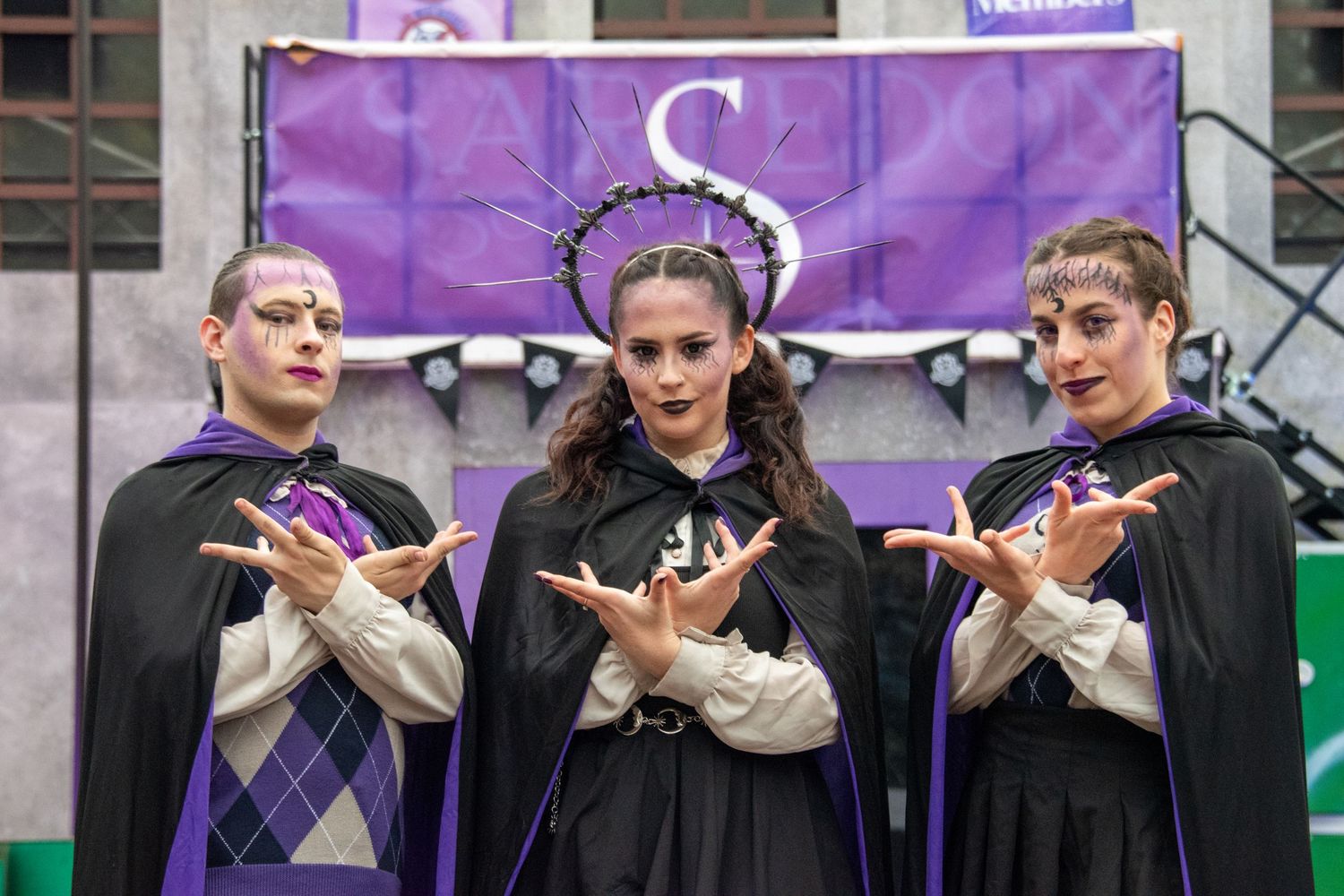 Three performers in matching purple and black capes with dramatic makeup pose on stage.
