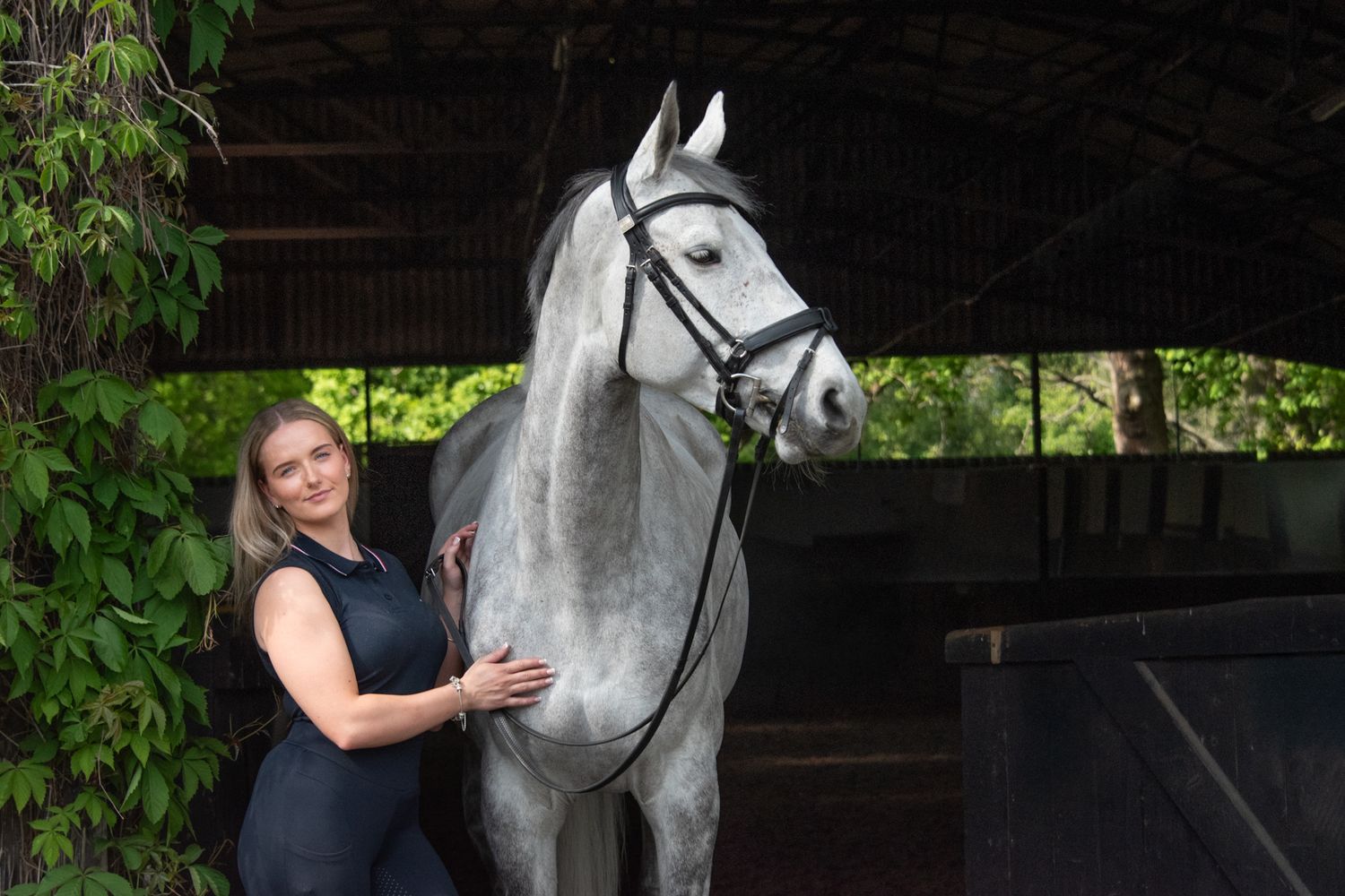 A smiling woman in a black dress stands beside a dapple gray horse in a stable yard surrounded by greenery.