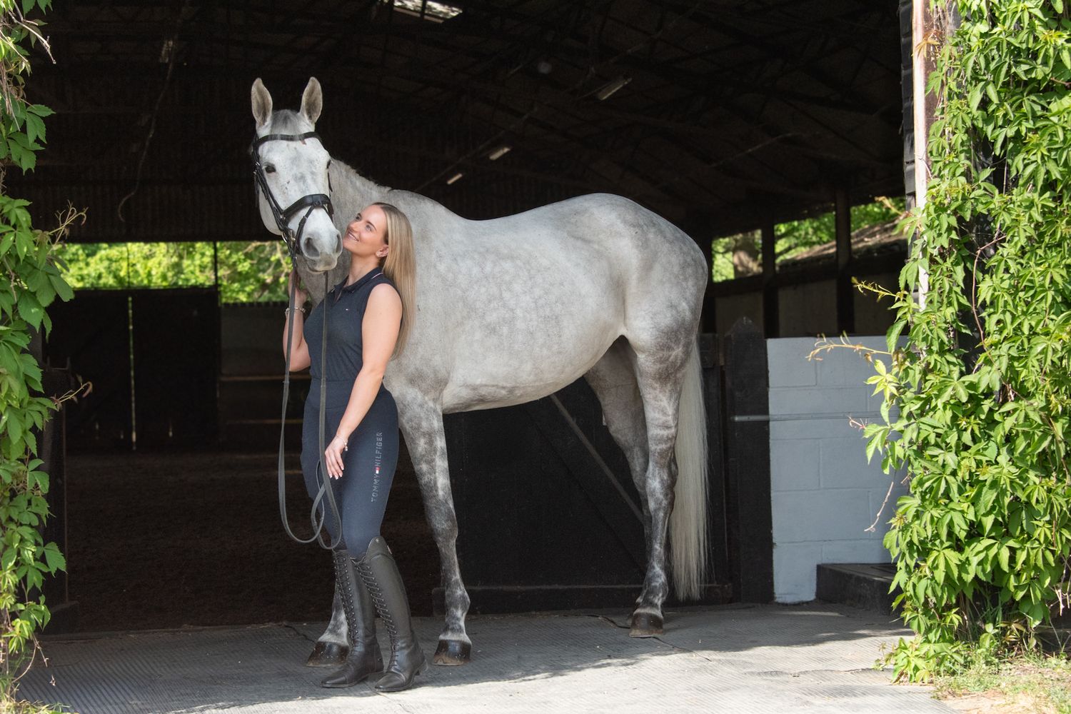 Woman standing with a grey horse in a stable barn doorway surrounded by green foliage.