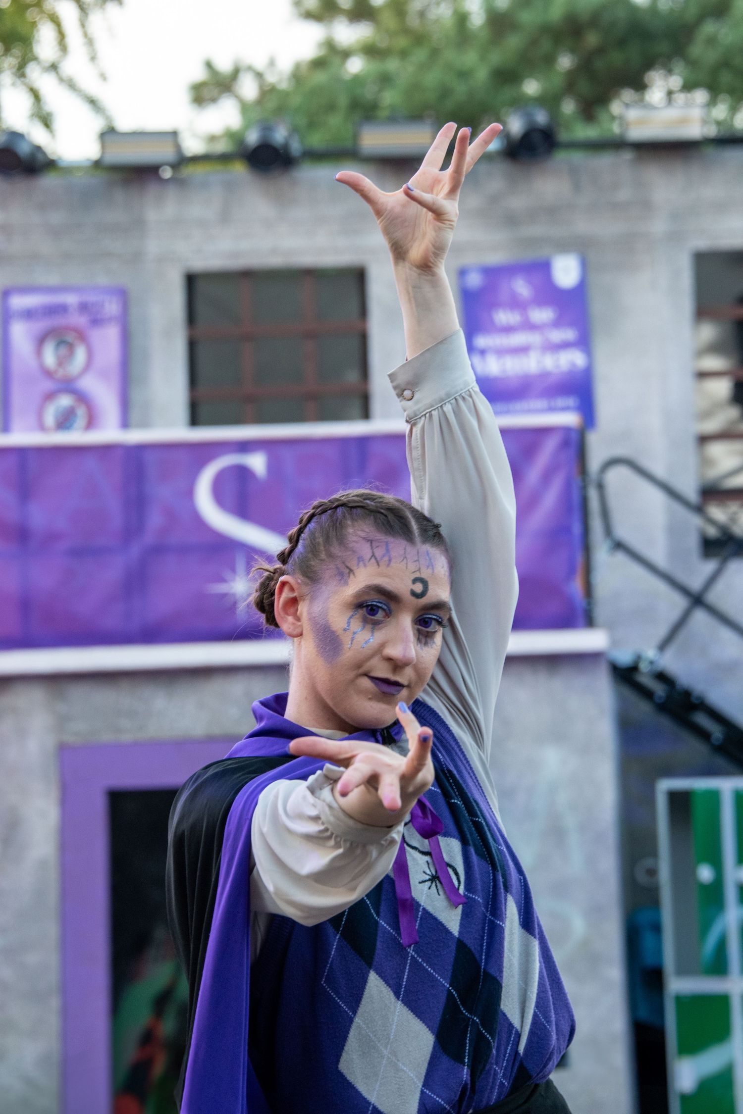 Performer with purple face paint and braided hair strikes a dramatic pose on outdoor stage.