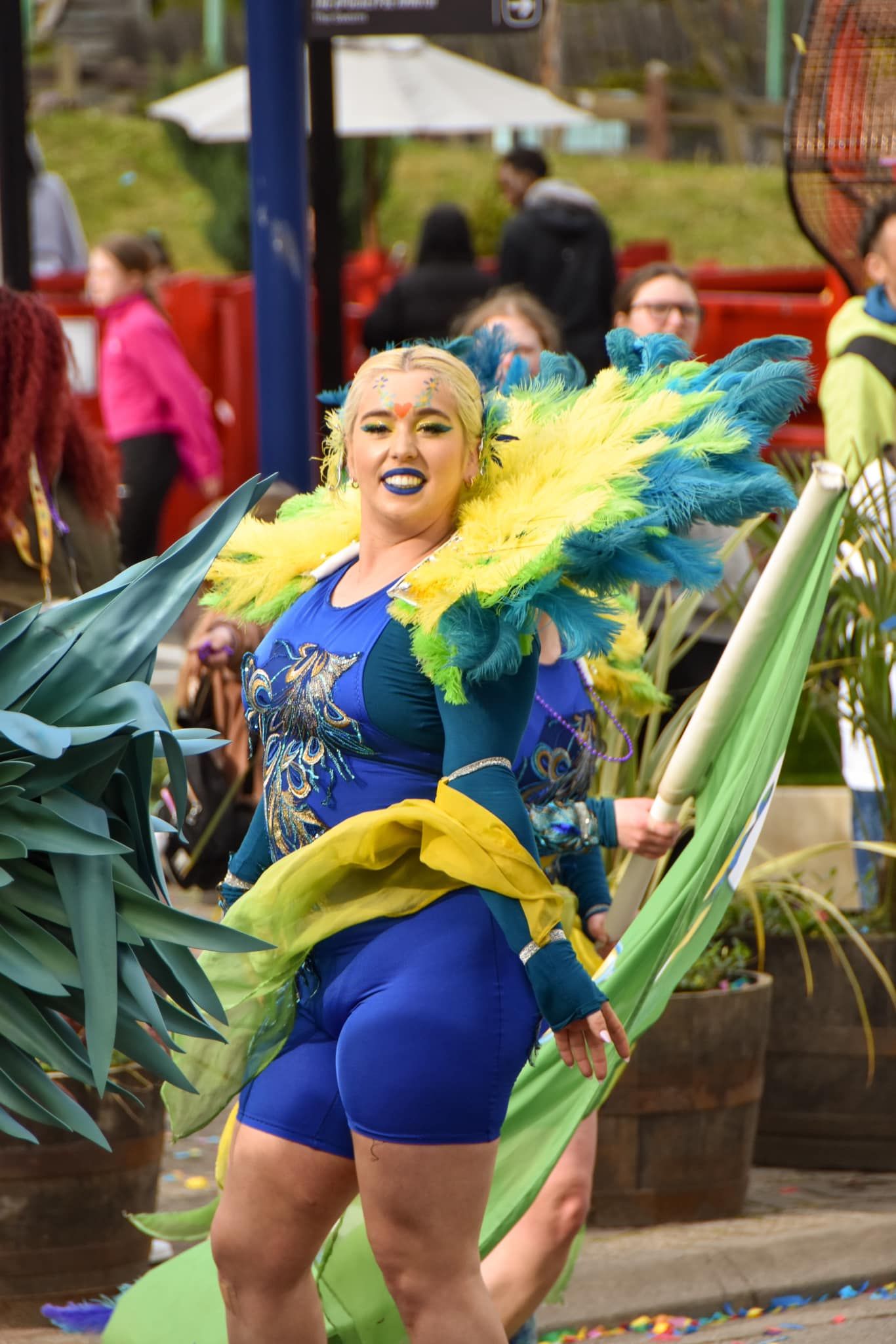 A performer in a blue outfit with yellow and teal feather accessories dances at a colorful carnival event.
