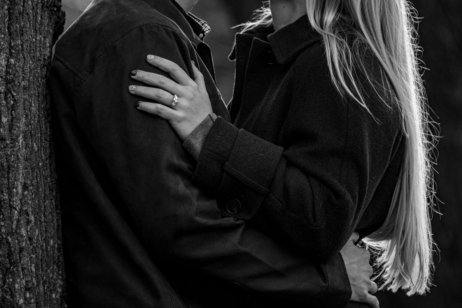 Black and white romantic photo of an engaged couple embracing by a tree, engagement ring visible.