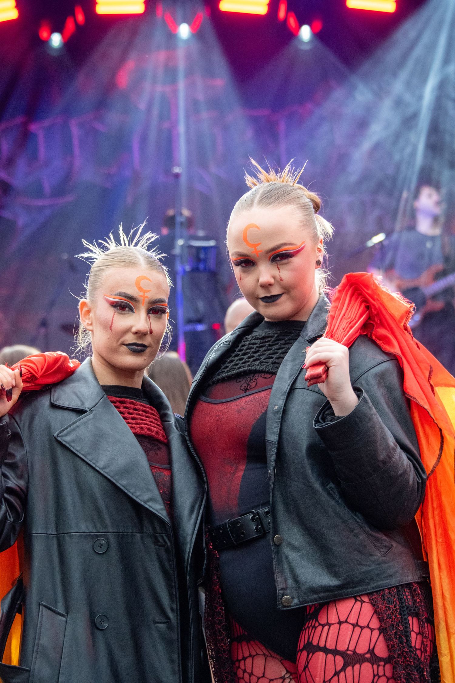 Two women in Halloween costumes with dramatic makeup and spiked hair posing at outdoor festival.