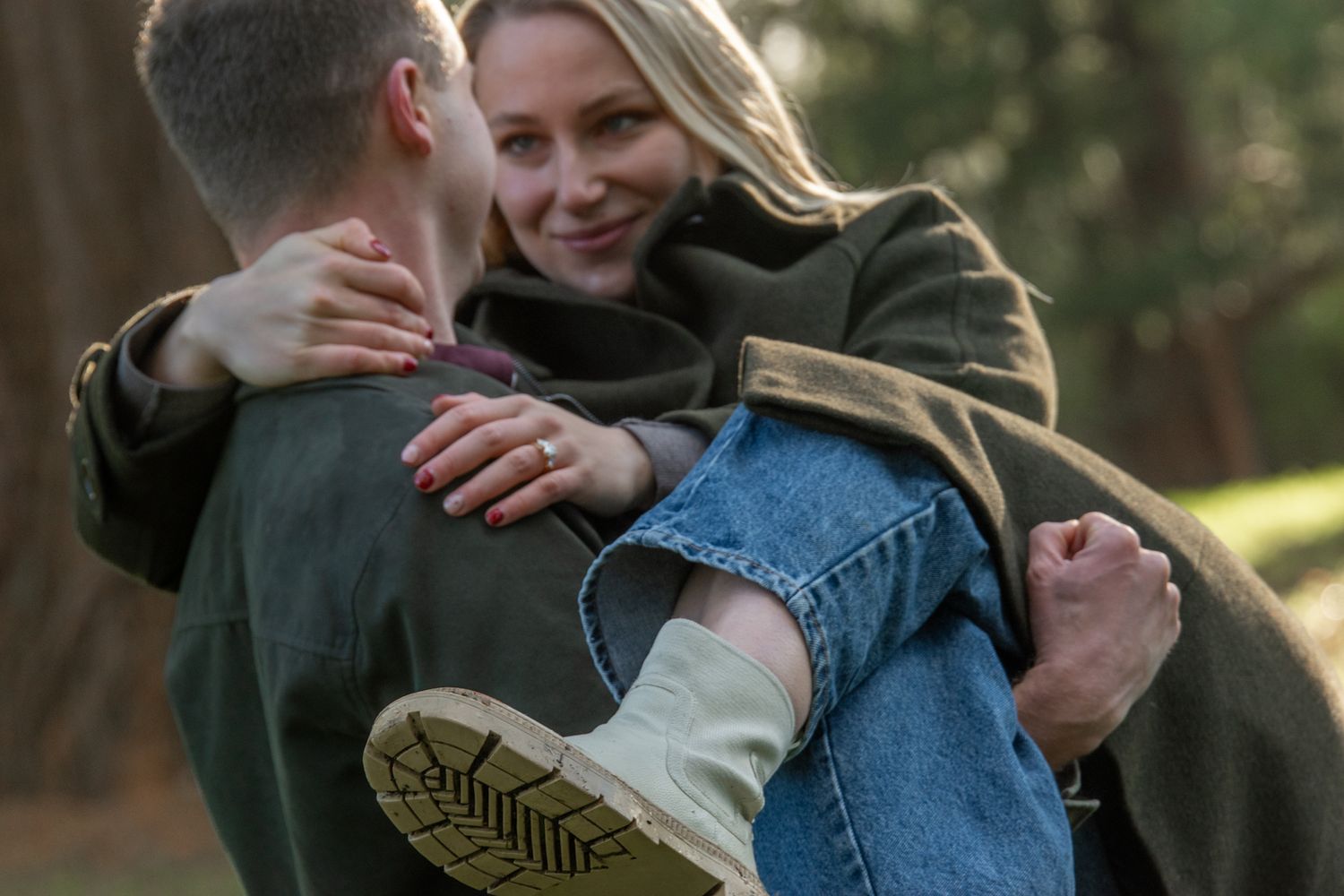 Man carrying smiling blonde woman wearing engagement ring in an outdoor setting.