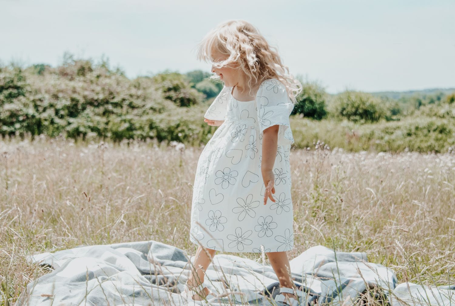 Young girl in a floral white dress standing on a blanket in a sunny meadow field.