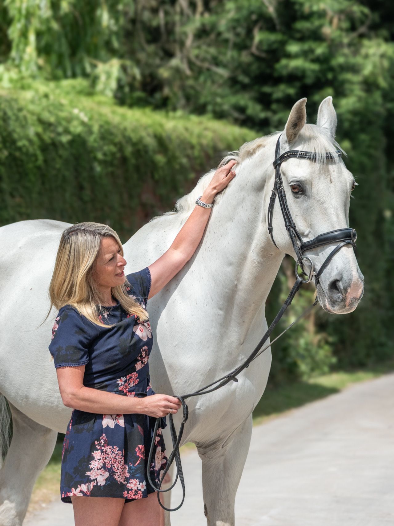 A blonde woman in a floral dress gently strokes a white horse wearing a bridle outdoors.