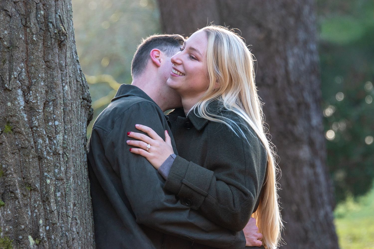 Romantic engaged couple sharing a kiss against a tree in a sunlit autumn park setting.