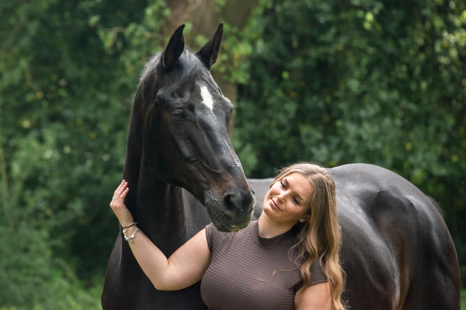 Young woman with blonde hair stands beside a black horse with a white blaze in a lush green setting.