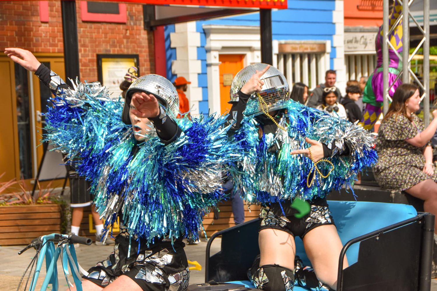 Two performers in silver helmets and blue tinsel costumes riding on a decorated carnival float.