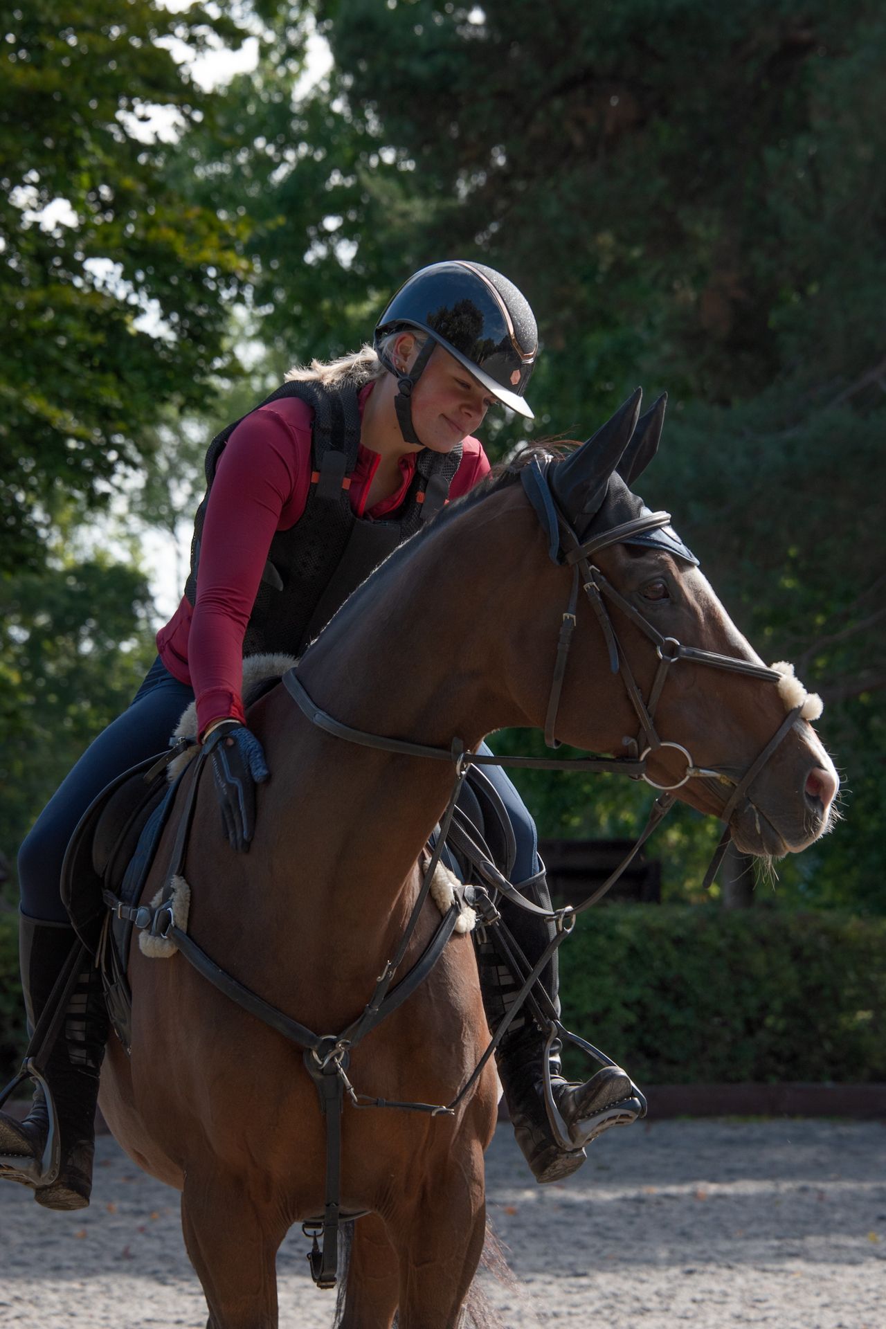 Female equestrian in a red shirt and helmet smiling while riding a bay horse in an outdoor arena.