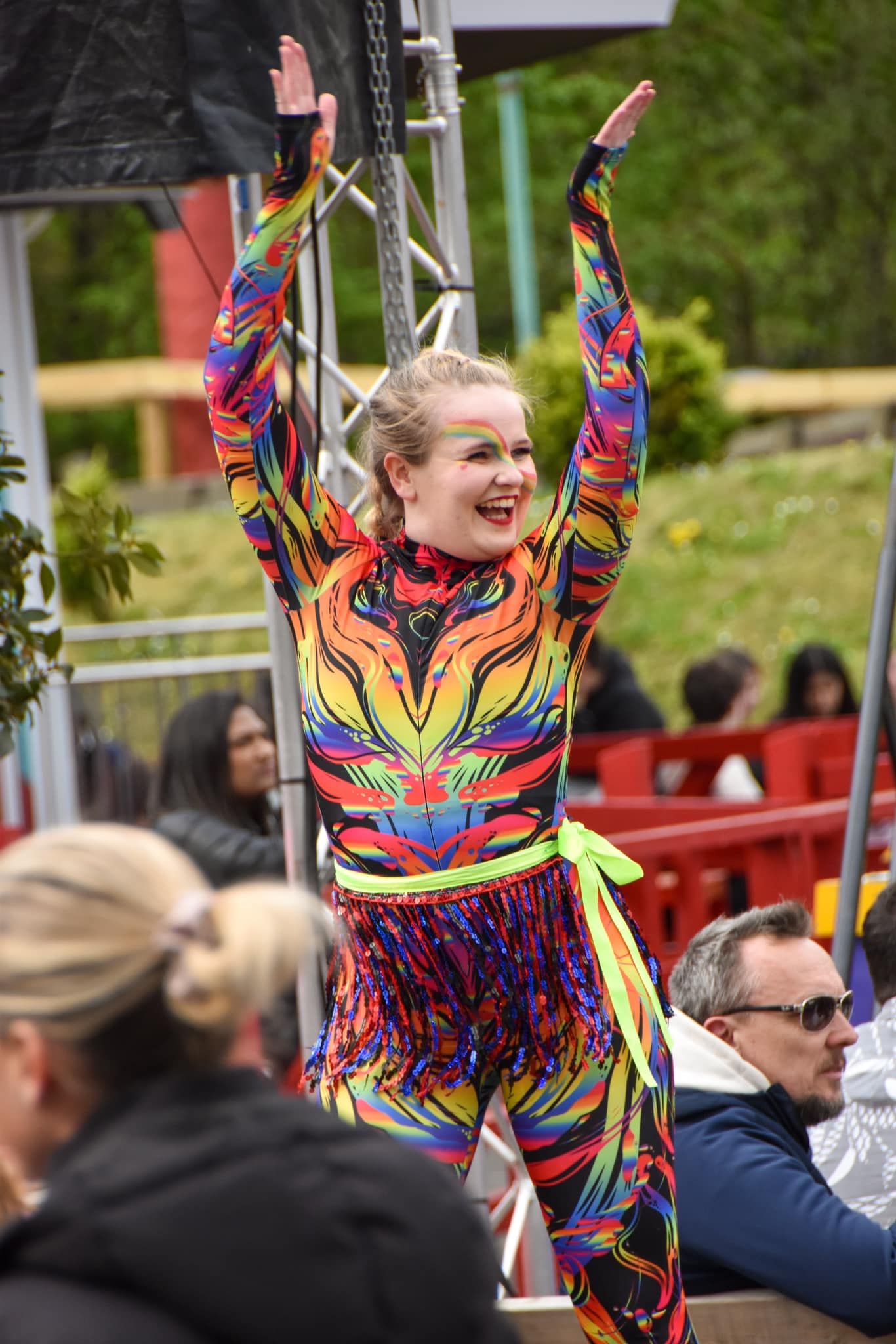 Energetic female performer in a rainbow-colored bodysuit raising her arms at an outdoor festival stage.
