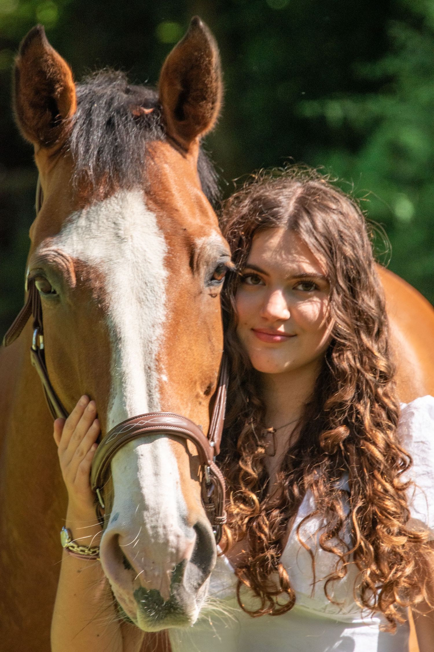 Young woman with long curly hair posing closely beside a chestnut horse with a white blaze in a sunny setting.