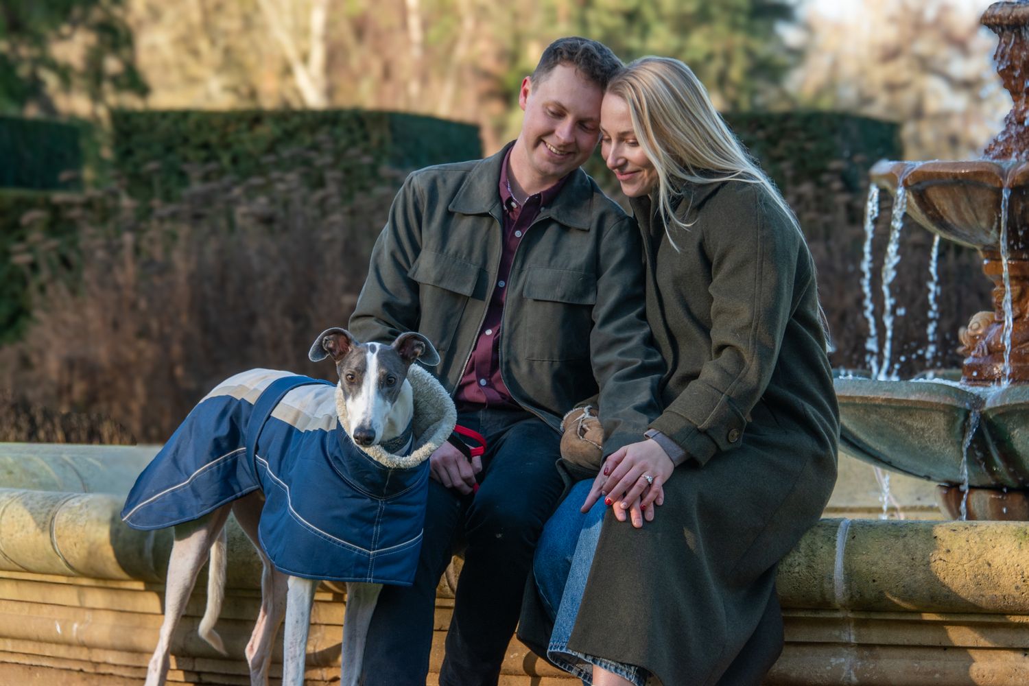 Engaged couple sitting by a fountain with their greyhound dog in a winter garden.