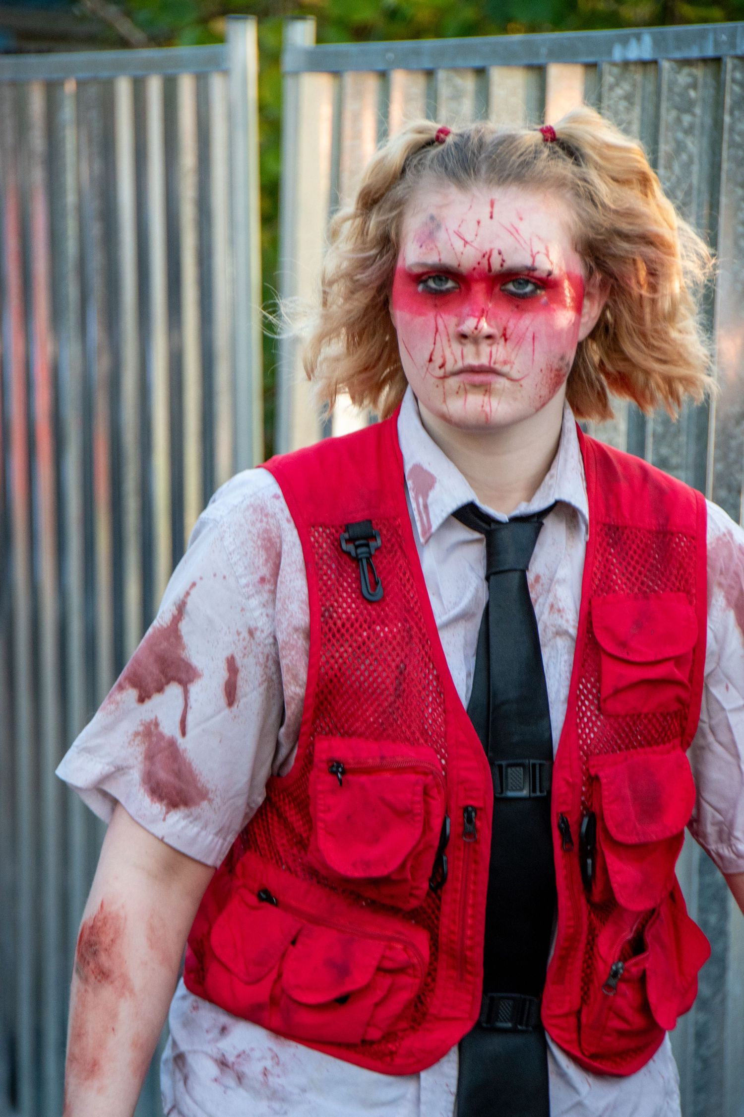 Young person in zombie Halloween costume with bloody face makeup, red vest, white shirt, and black tie.