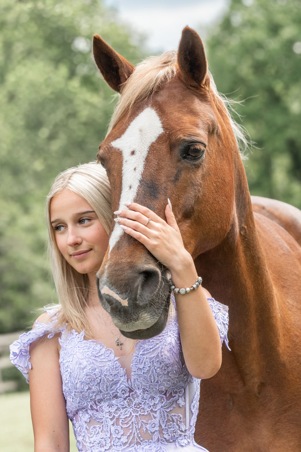 Young blonde woman in lavender lace dress gently holds face of chestnut horse with white blaze outdoors.