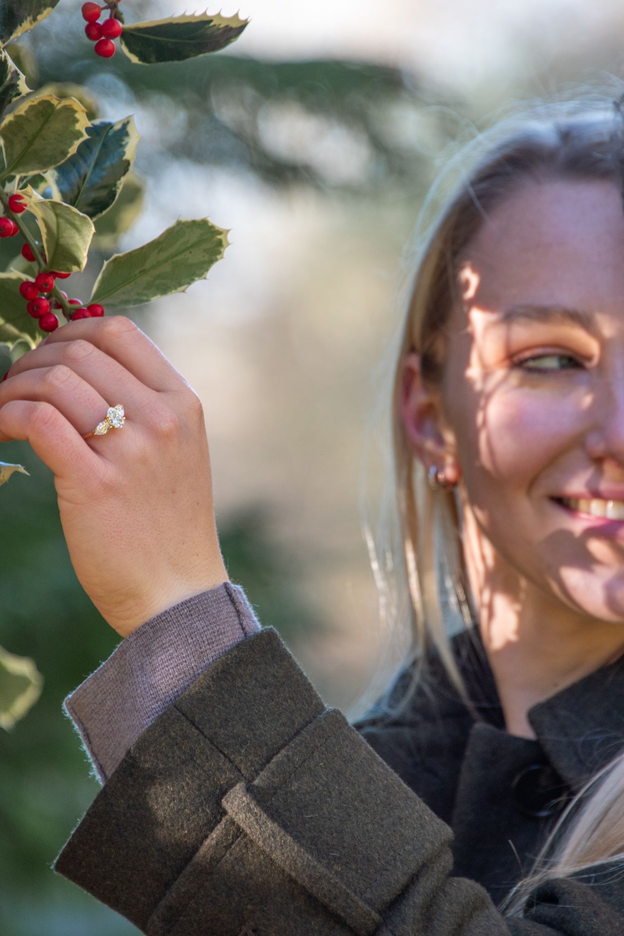 Smiling blonde woman wearing engagement ring touching holly berries in sunlight.
