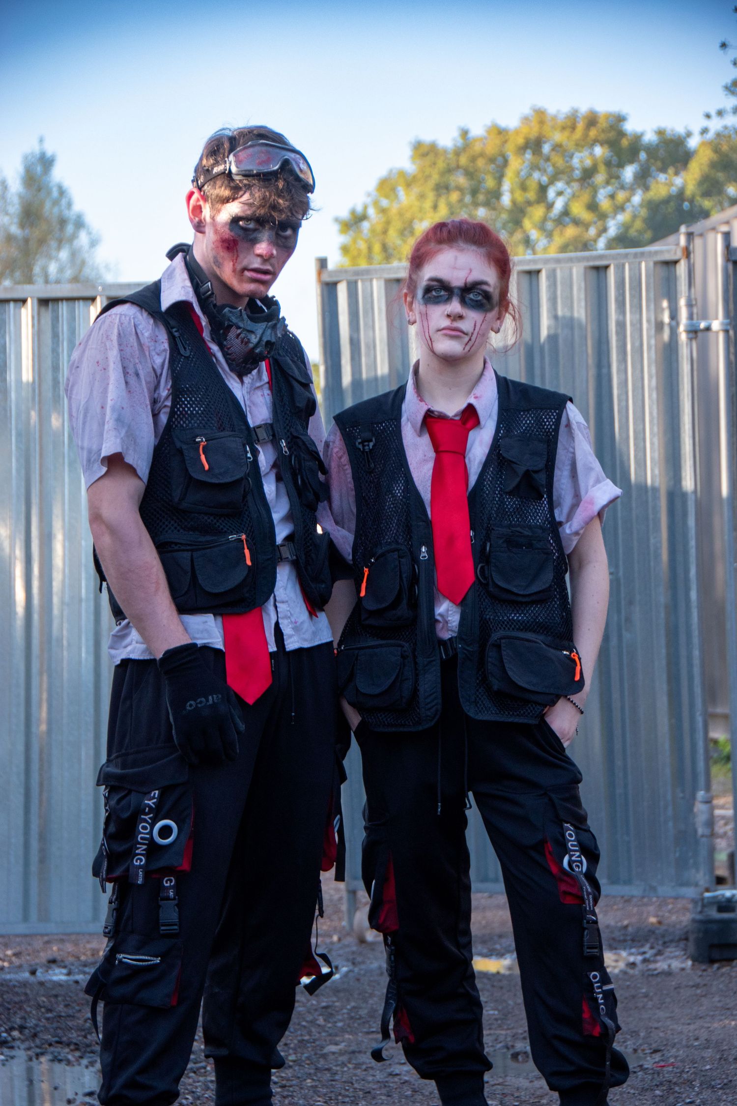 Two teens in zombie makeup wearing black tactical vests and red ties at Halloween event.