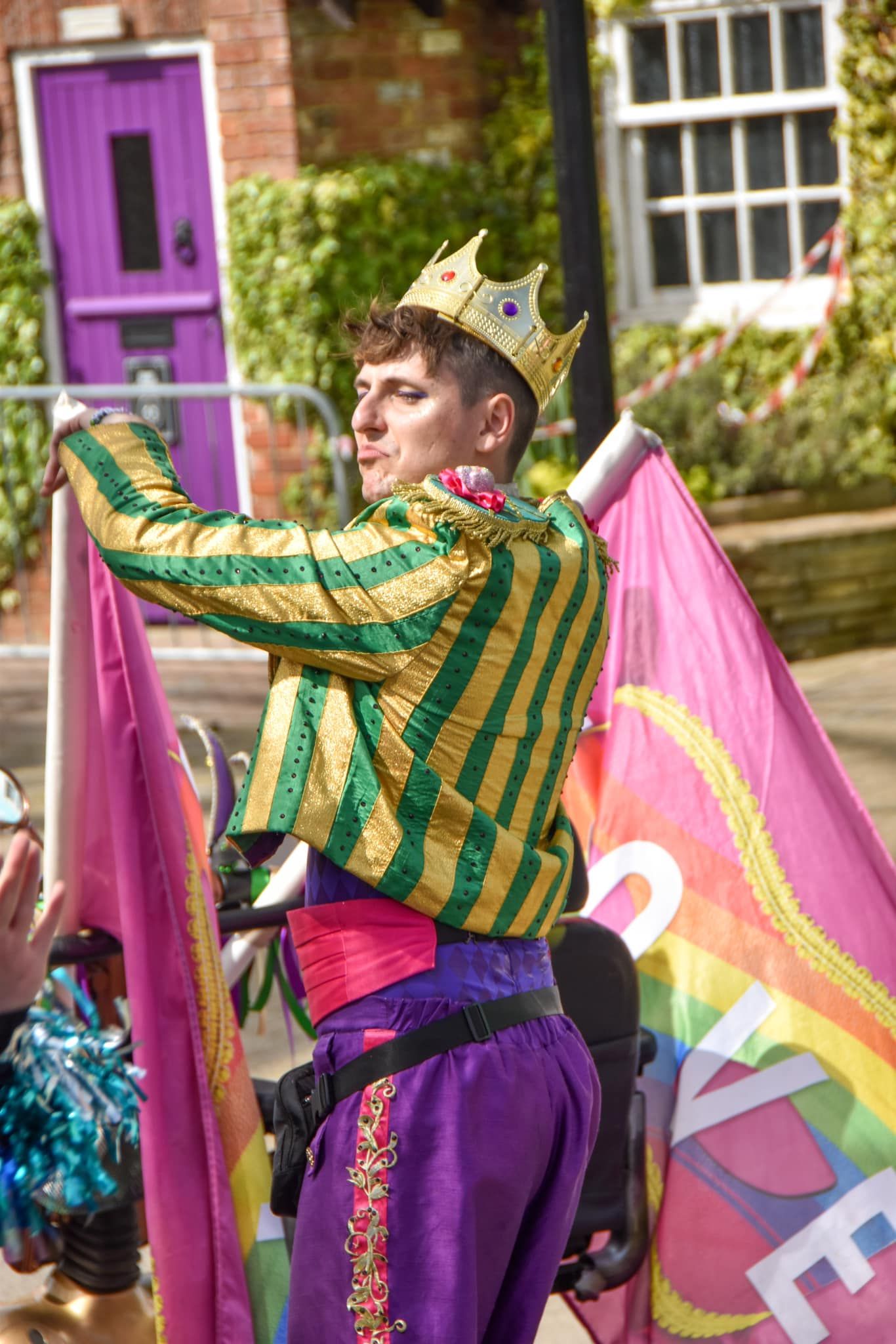 A performer in a gold and green striped costume with a crown participates in a colorful Pride parade.