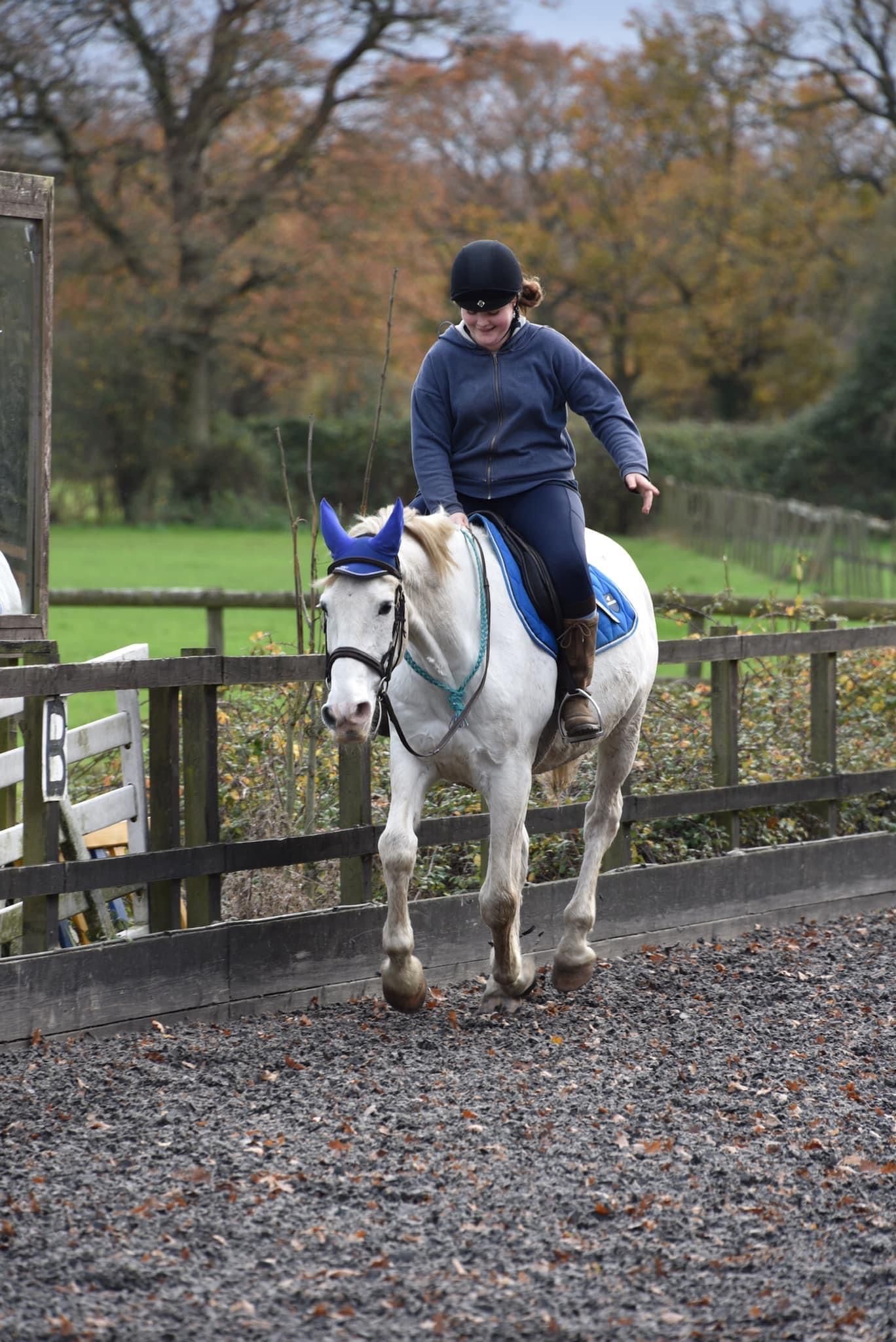 Female rider in navy outfit on a white grey pony trotting in an outdoor arena with autumn trees in background.