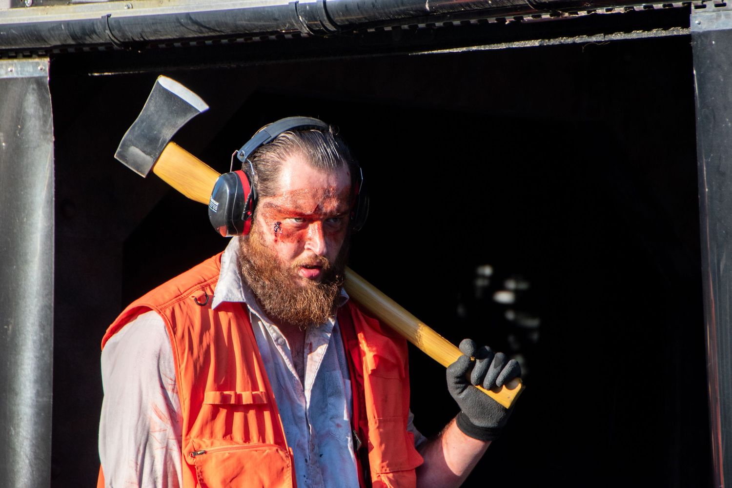Bearded man in horror costume with bloody face paint, orange vest, headphones, and axe prop.