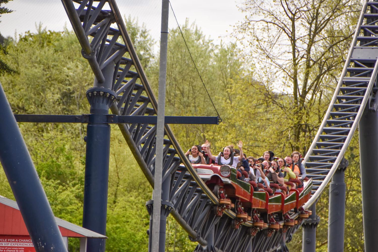 Long train of roller coaster riders descending a wooden track surrounded by trees at a theme park.