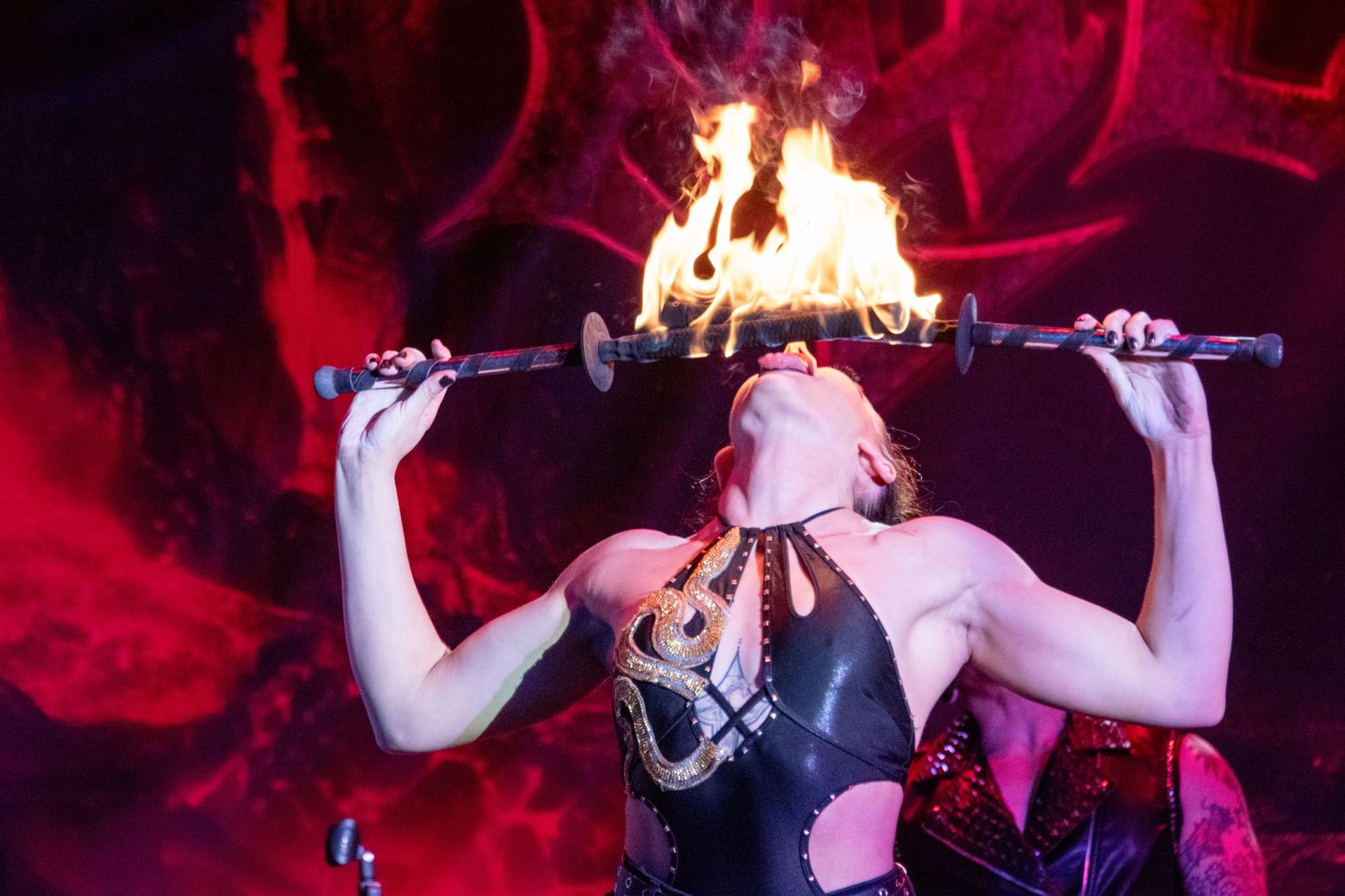 Performer balancing a flaming torch in mouth during a dramatic fire-eating act under red stage lighting.