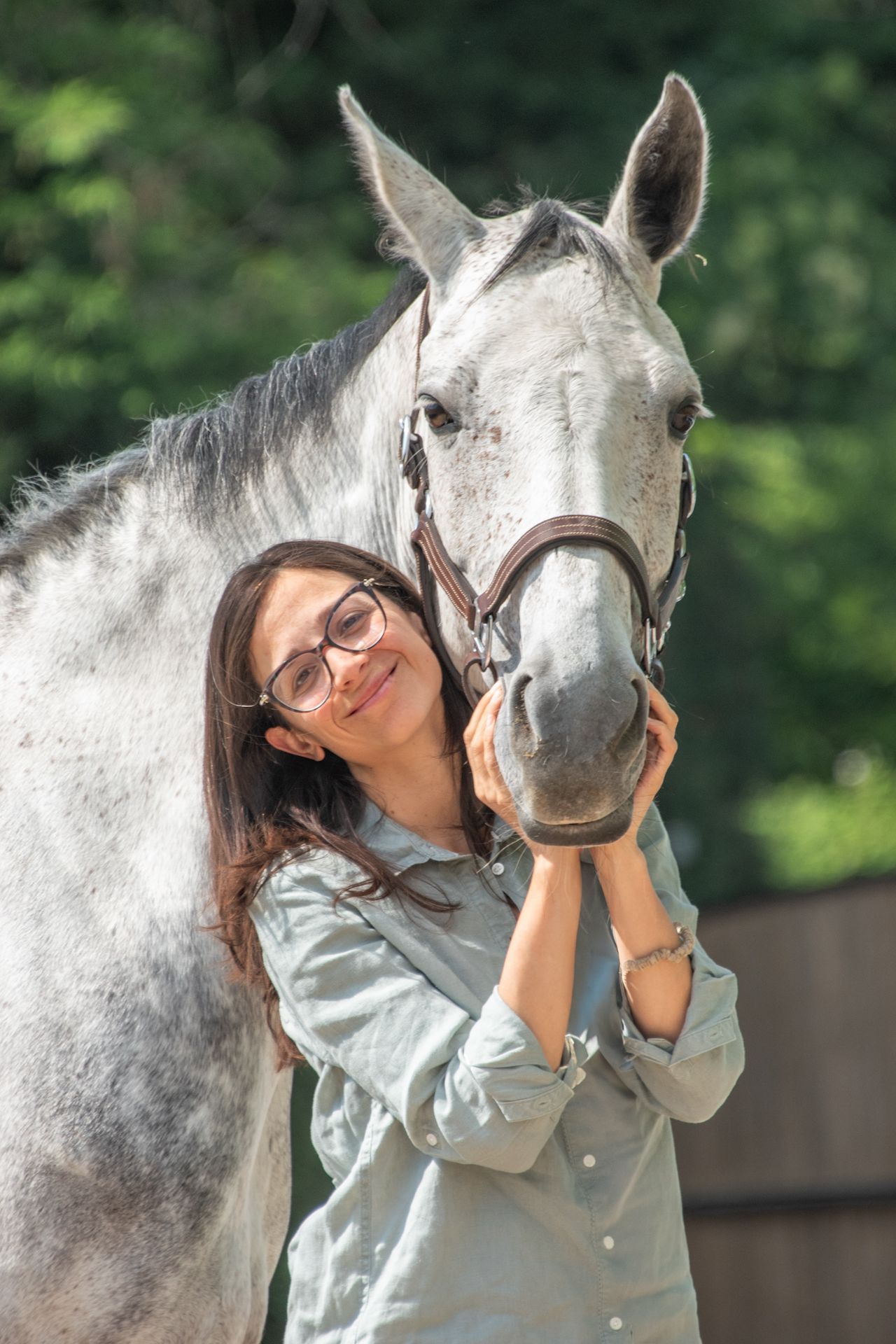 A smiling woman with glasses gently holds the face of a grey horse in a sunny outdoor setting.